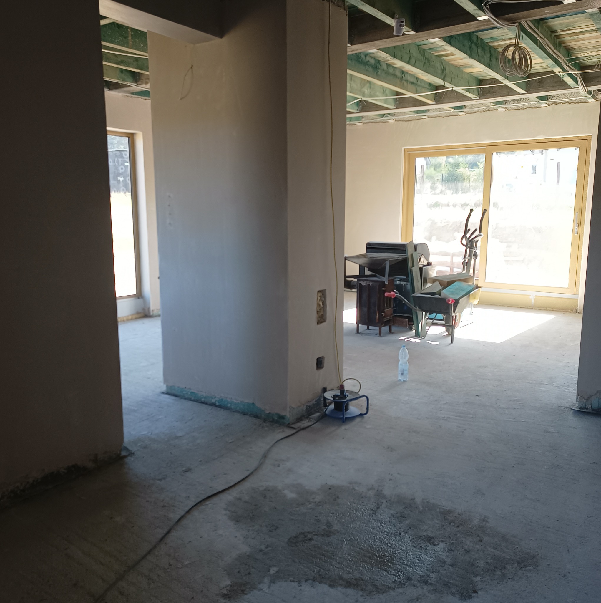 Wnętrze surowego domu w trakcie prac wykończeniowych: świeżo otynkowane ściany, widoczny strop, wózek budowlany i butelka wody na podłodze, kabel od urządzenia podłączony do ściany.