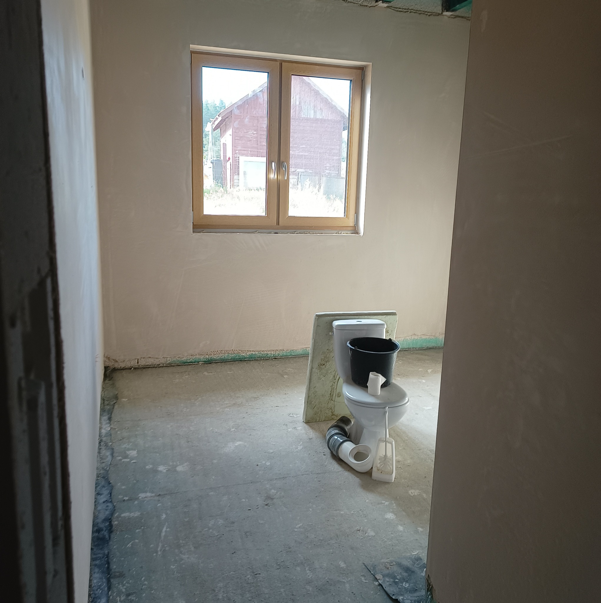 Wnętrze w stanie surowym po nałożeniu gładzi gipsowej na ścianach, widoczne okno z drewnianą ramą, prowizoryczna toaleta z wiadrem i szczotką.