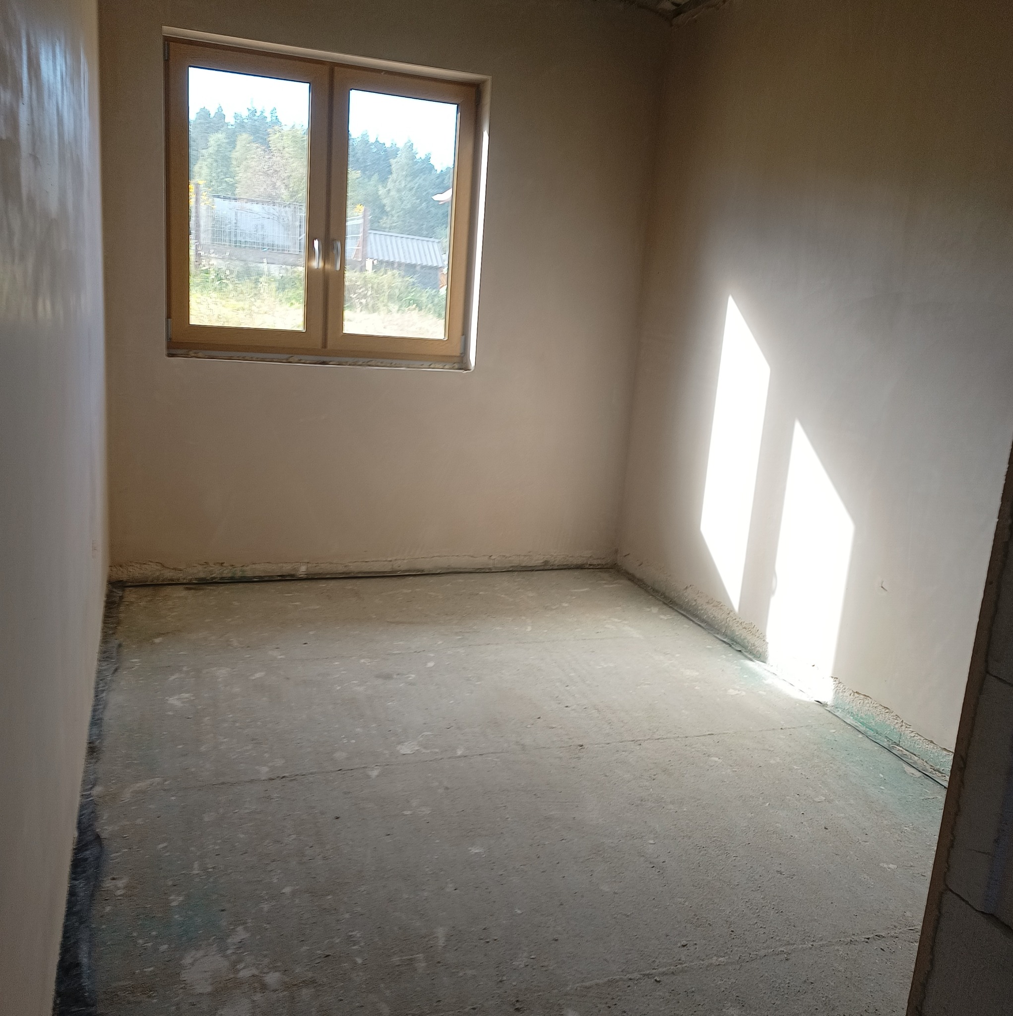 Wnętrze pokoju w trakcie prac wykończeniowych, ściany pokryte świeżym tynkiem, widoczne zacieki i nierówności, okno z widokiem na zieleń na zewnątrz, podłoga surowa, przygotowana do dalszych prac.