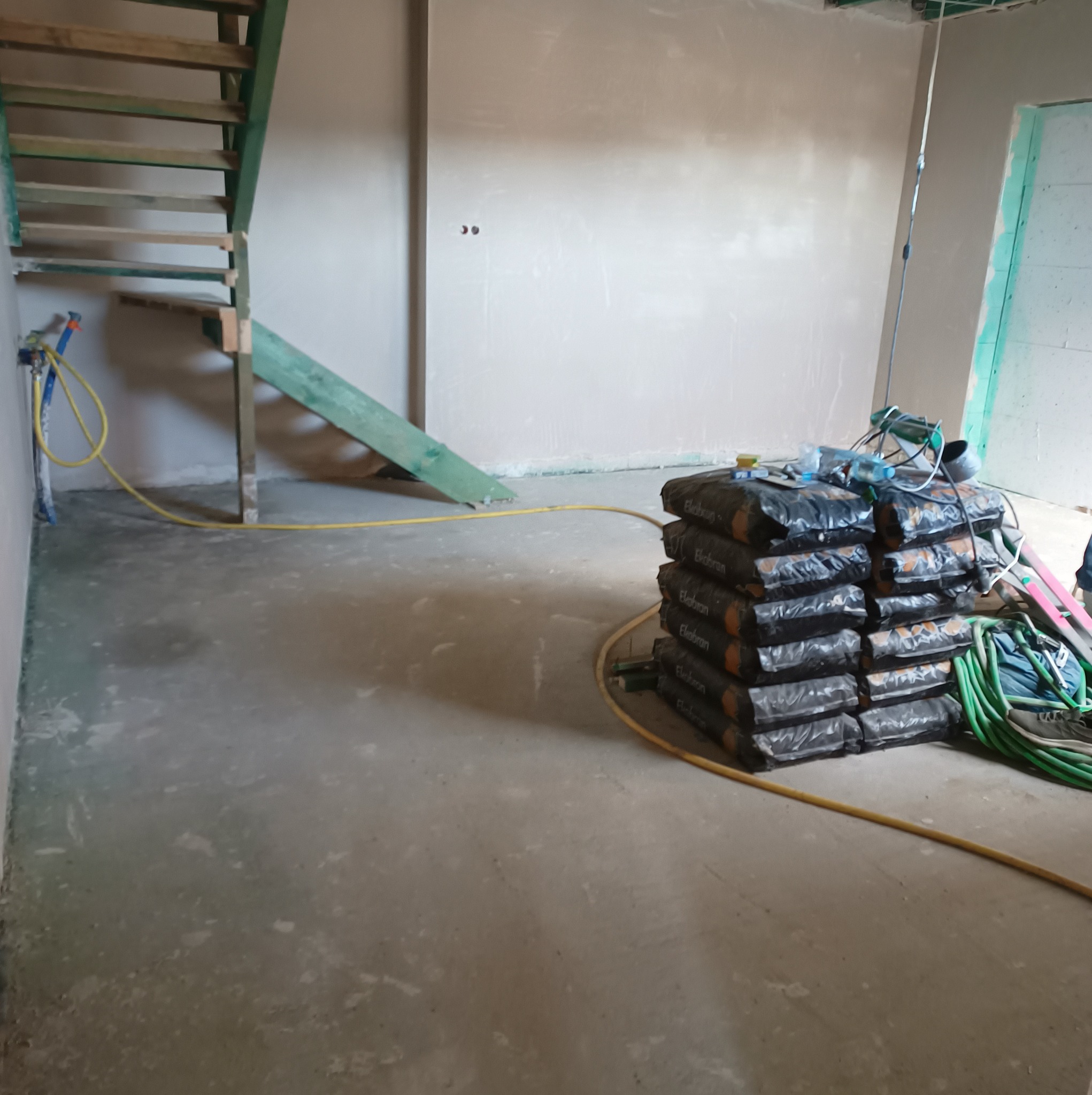 Wnętrze w trakcie prac wykończeniowych: ściany po nałożeniu gładzi, sterta worków z zaprawą EkoBran przy ścianie, pod schodami węże i narzędzia.