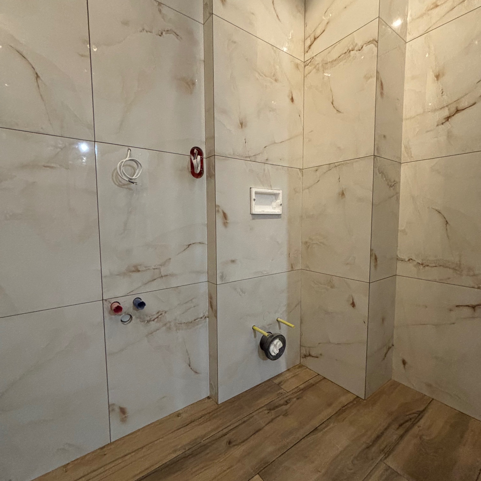 Fragment łazienki w trakcie wykańczania, widoczne płytki imitujące marmur na ścianach, drewnopodobna podłoga, przygotowane instalacje wodno-kanalizacyjne, oraz puszka podtynkowego zestawu WC.