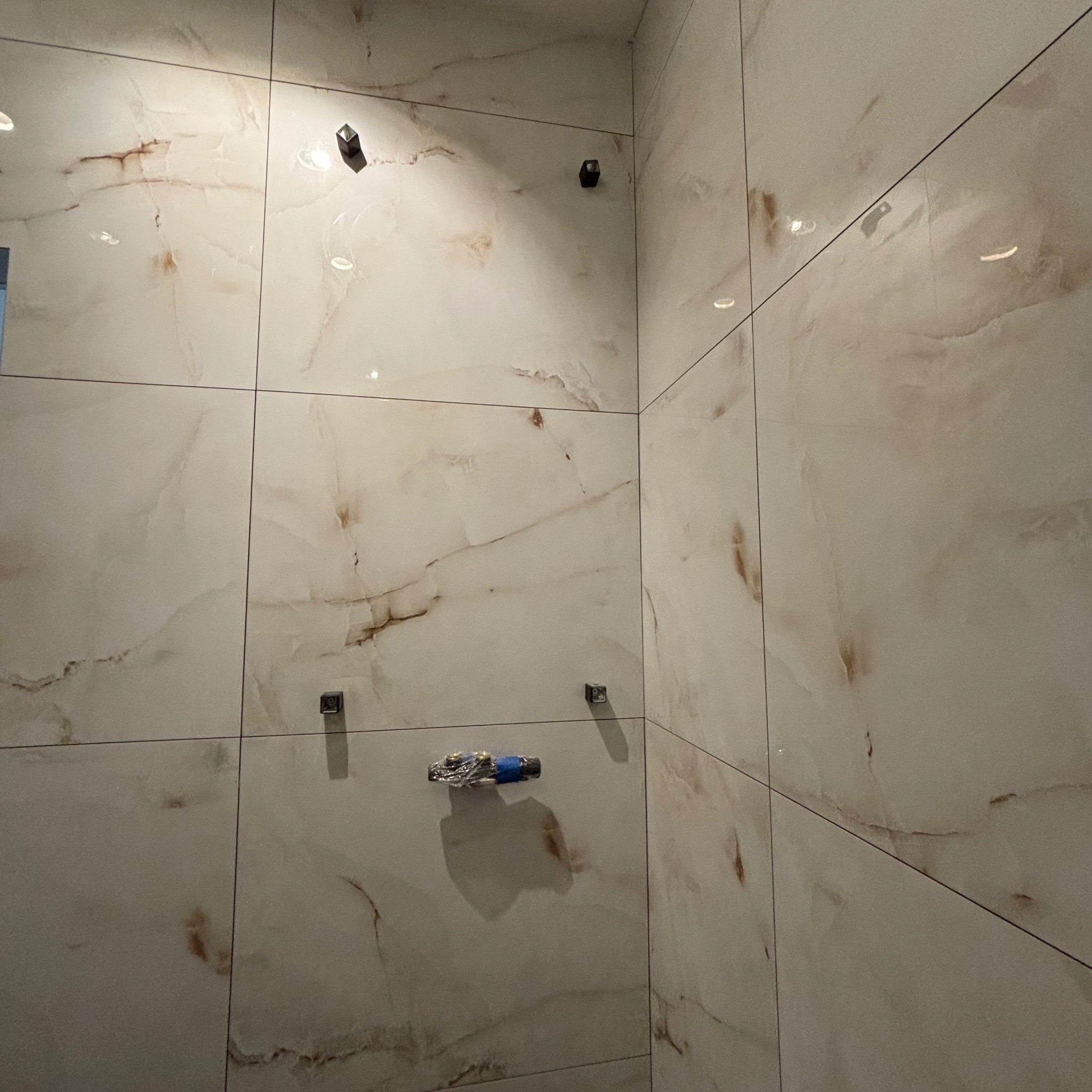 Ściana w łazience wyłożona dużymi, jasnymi płytkami imitującymi marmur, z zamontowanymi elementami armatury zabezpieczonymi folią i taśmą malarską.