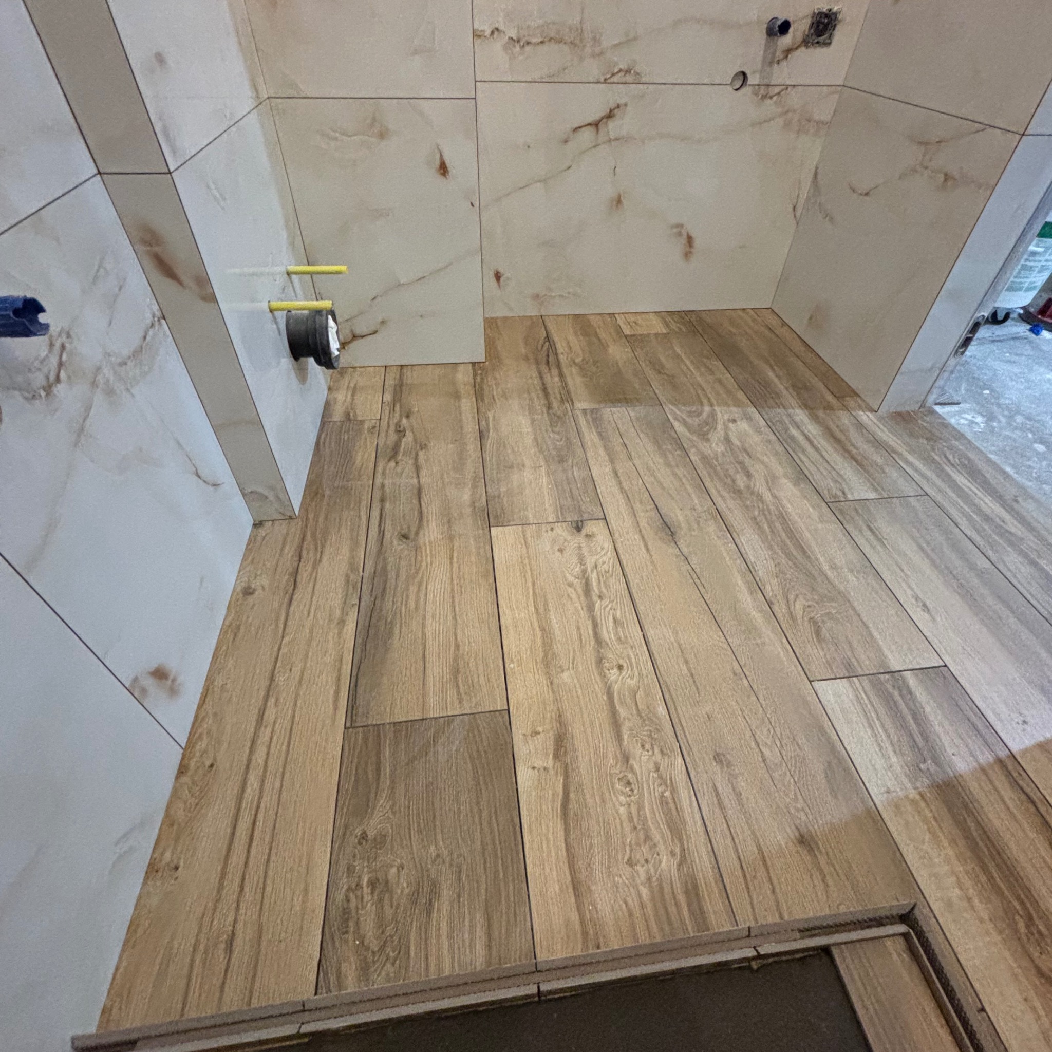 Ukończona łazienka z płytkami imitującymi drewno na podłodze i marmurowe płytki na ścianach, widoczne rury instalacyjne i otwór na gniazdko.