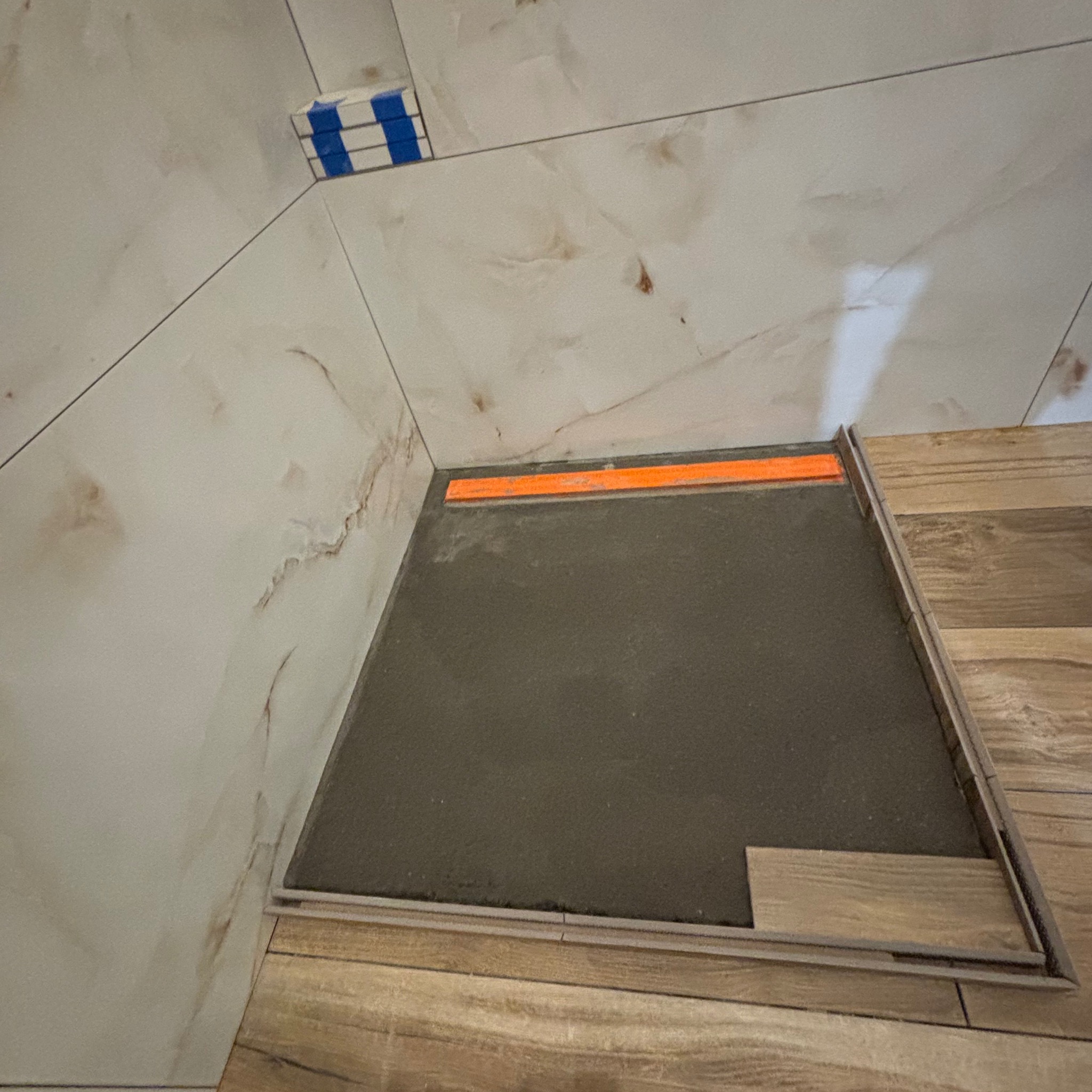 Przygotowanie do wylewki podłogowej w narożniku łazienki, widoczne płytki ścienne imitujące marmur, listwa poziomująca i częściowo ułożona podłoga drewnopodobna.