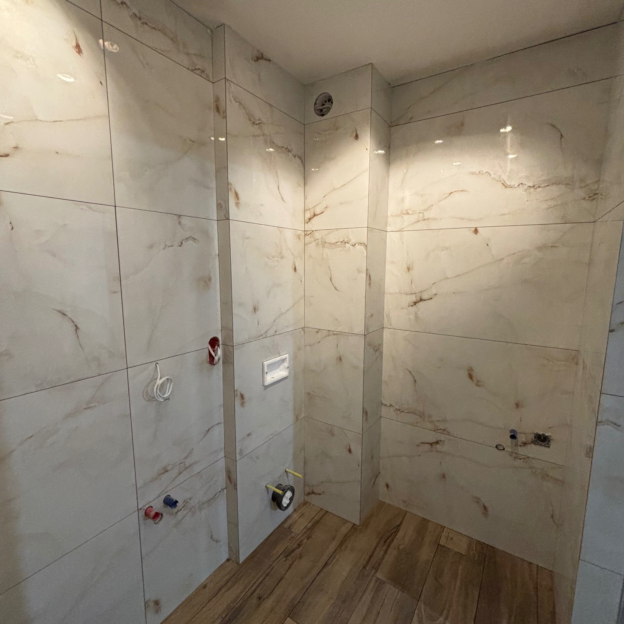 Wykończona łazienka z beżowymi, marmurkowymi płytkami na ścianach i drewnopodobną podłogą, widoczne wystające rury i instalacja sanitarna przed montażem armatury.