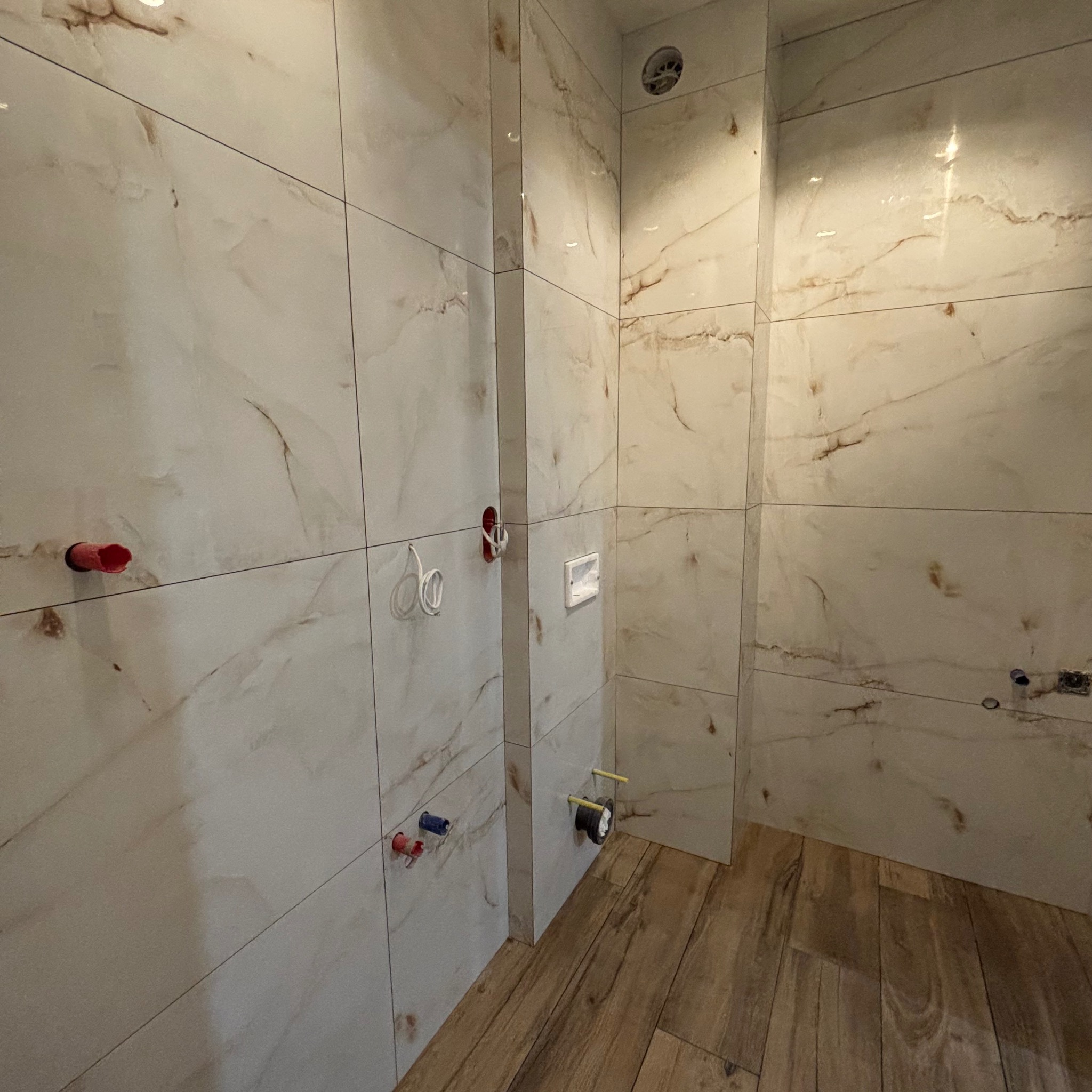 Wnętrze łazienki w trakcie remontu: ściany wyłożone płytkami imitującymi marmur, drewnopodobna podłoga, widoczne instalacje wodno-kanalizacyjne przed montażem armatury.