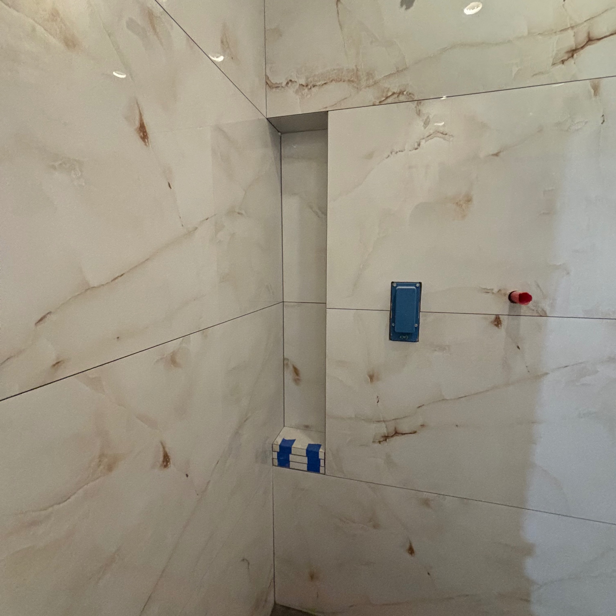 Ściana wyłożona płytkami imitującymi marmur z wnęką w rogu, zabezpieczoną niebieską taśmą malarską, widoczna puszka elektryczna i wystający fragment rurki.
