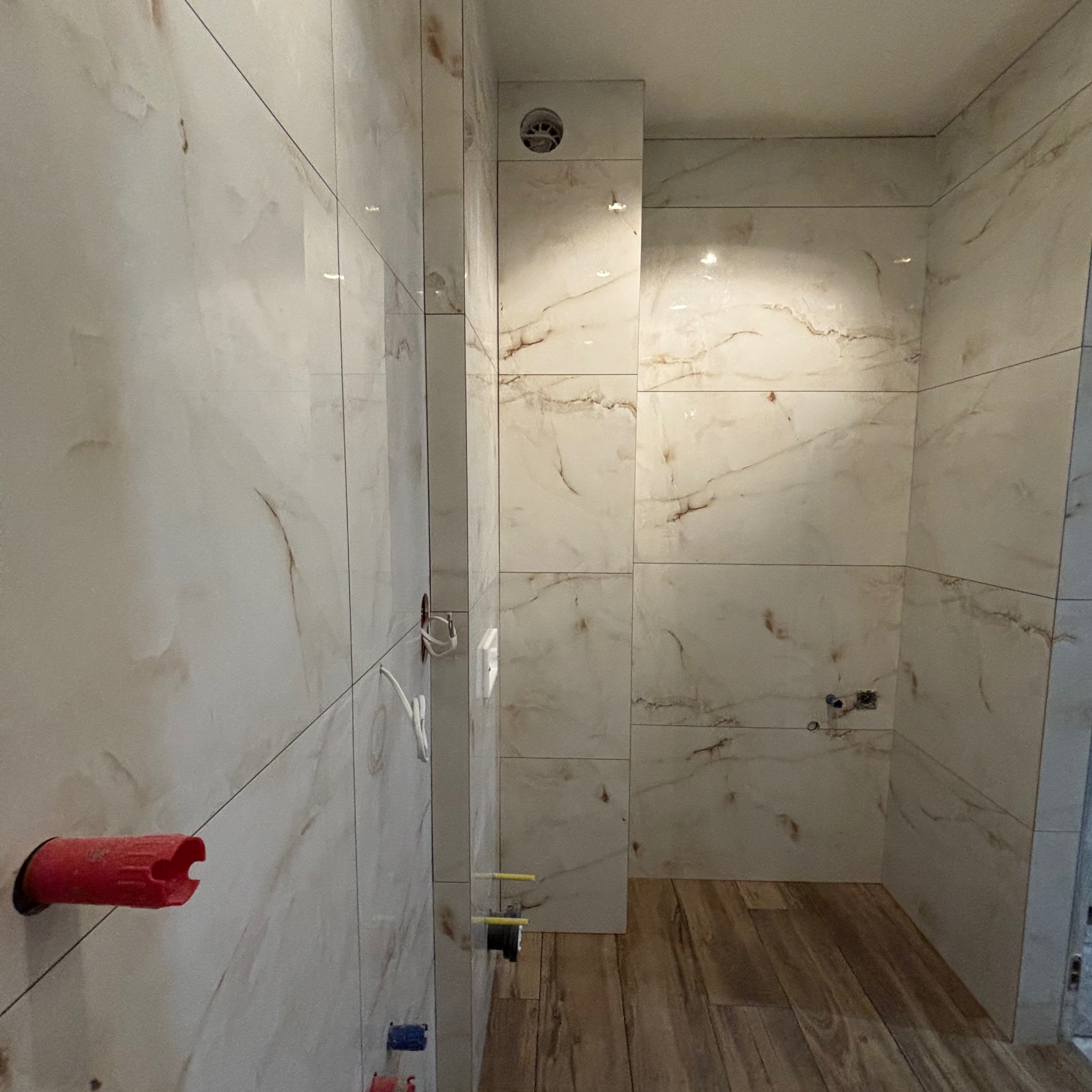 Fragment łazienki w trakcie wykańczania, widoczne płytki imitujące marmur na ścianach, drewnopodobna podłoga, wystające rury i kable w ścianach, przygotowane pod instalacje.