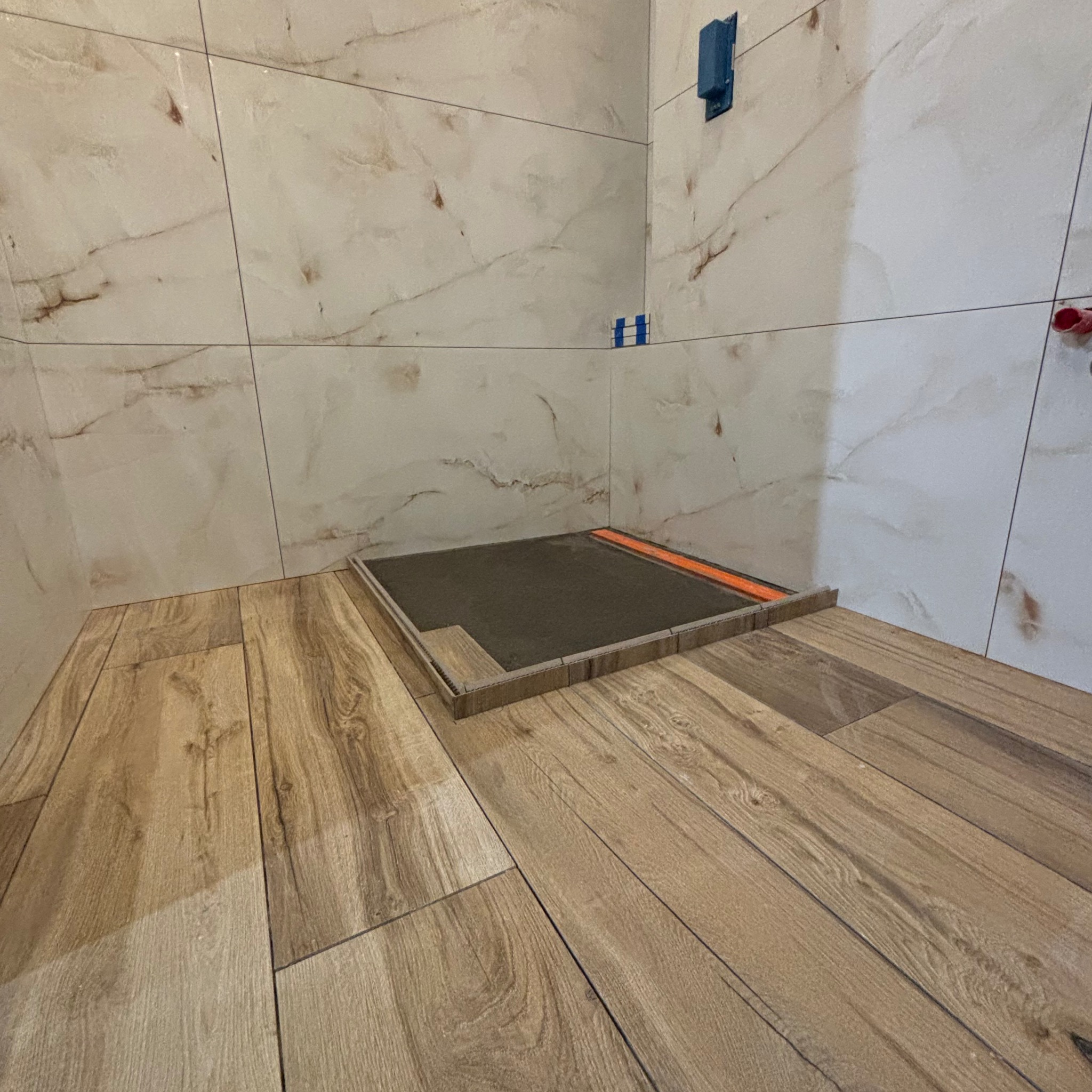 Przygotowanie do montażu odpływu liniowego w kabinie prysznicowej, widoczne płytki imitujące drewno na podłodze i marmurowe płytki na ścianach.