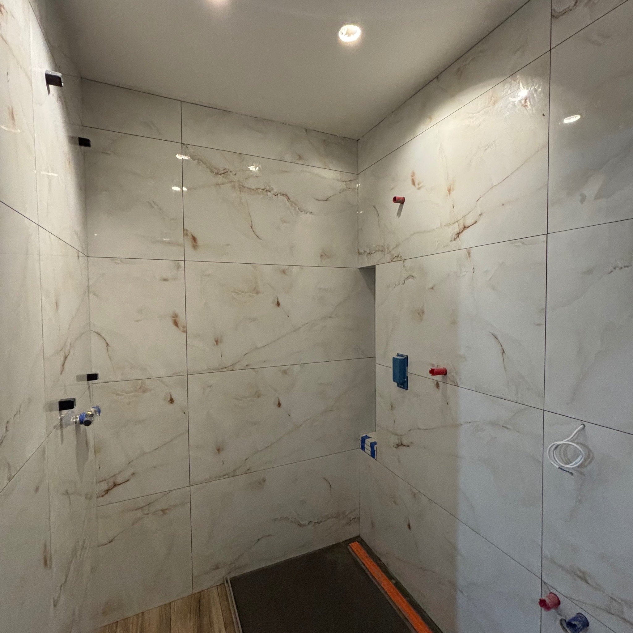 Świeżo wykafelkowana ściana w kabinie prysznicowej z imitacją marmuru, widoczne przygotowane wyprowadzenia wodne i elektryczne, oraz zamontowany odpływ liniowy.