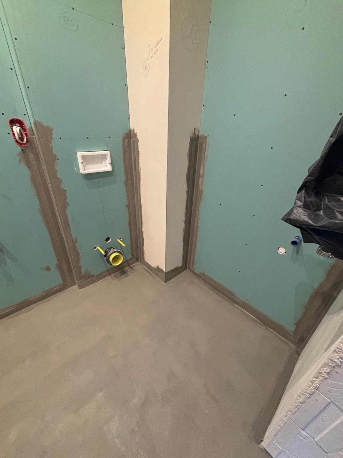 Wnętrze łazienki w trakcie remontu: zielone płyty na ścianach, szpachlowane łączenia, widoczne instalacje wodno-kanalizacyjne z żółtymi elementami, wylewka podłogowa, narożnik z kolumną.