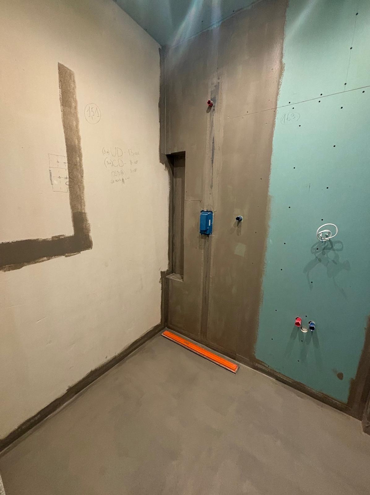 Łazienka w trakcie remontu: częściowo położona płyta karton gips, fragmenty ścian z gładzią, widoczne instalacje wodne i elektryczne, poziomica na podłodze i notatki na ścianie.