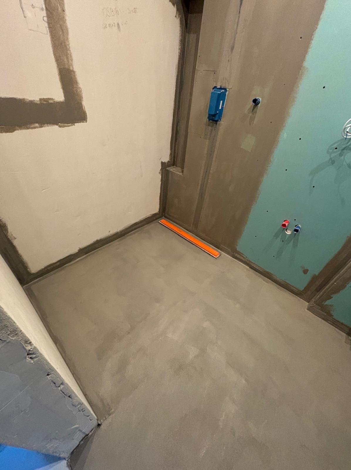 Przygotowanie podłoża w łazience: wylewka samopoziomująca na podłodze, ściany częściowo pokryte gruntem, widoczna poziomica i instalacja wodna.