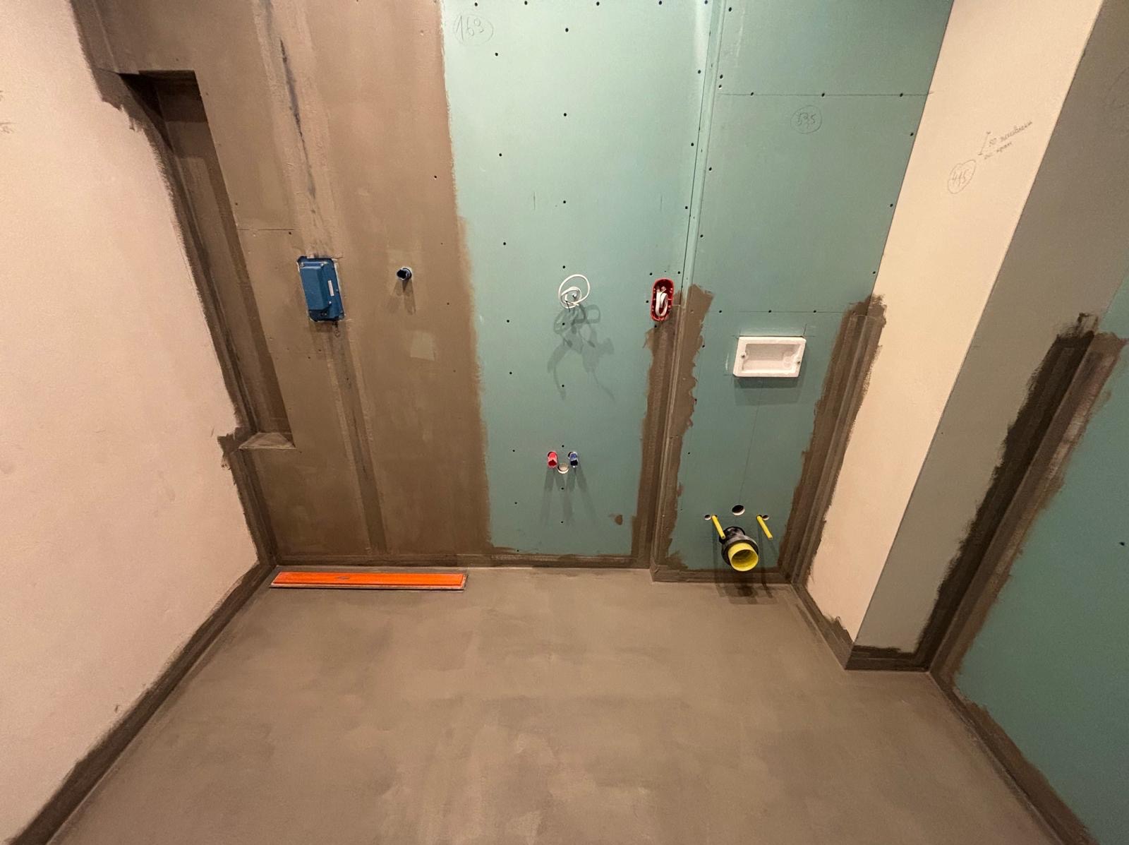 Łazienka w trakcie remontu: surowy beton na podłodze, częściowo położona płyta kartonowo-gipsowa na ścianach z widocznymi instalacjami elektrycznymi i hydraulicznymi. Na podłodze leży poziomica.