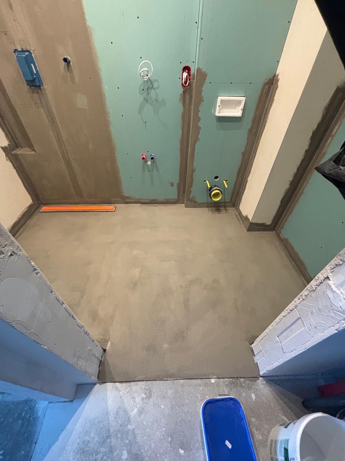 Przygotowane do wykończenia wnętrze łazienki z zamontowaną płytą kartonowo-gipsową na ścianach i suficie, świeżo wylaną wylewką podłogową, widocznymi instalacjami hydraulicznymi i elektrycznymi...