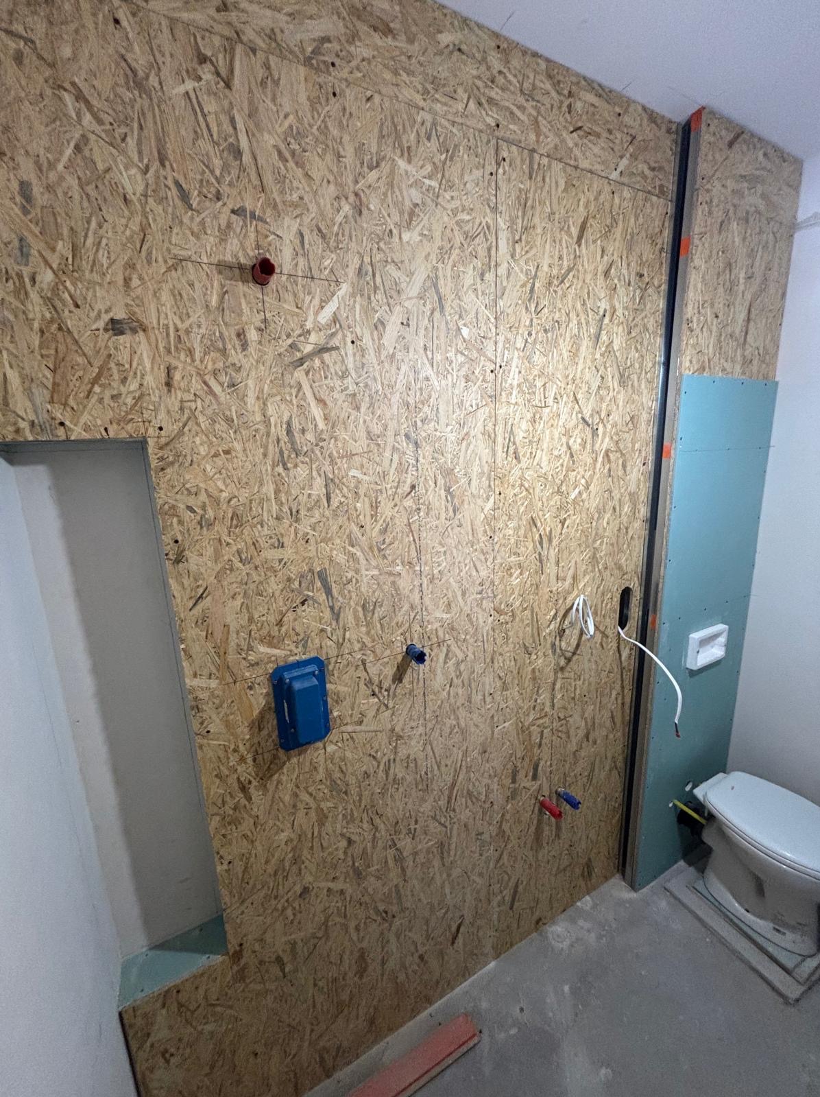 Wnętrze łazienki w trakcie prac wykończeniowych, ściana obłożona płytami OSB z widocznymi instalacjami wodnymi i elektrycznymi, częściowo zamontowana płyta gipsowo-kartonowa.