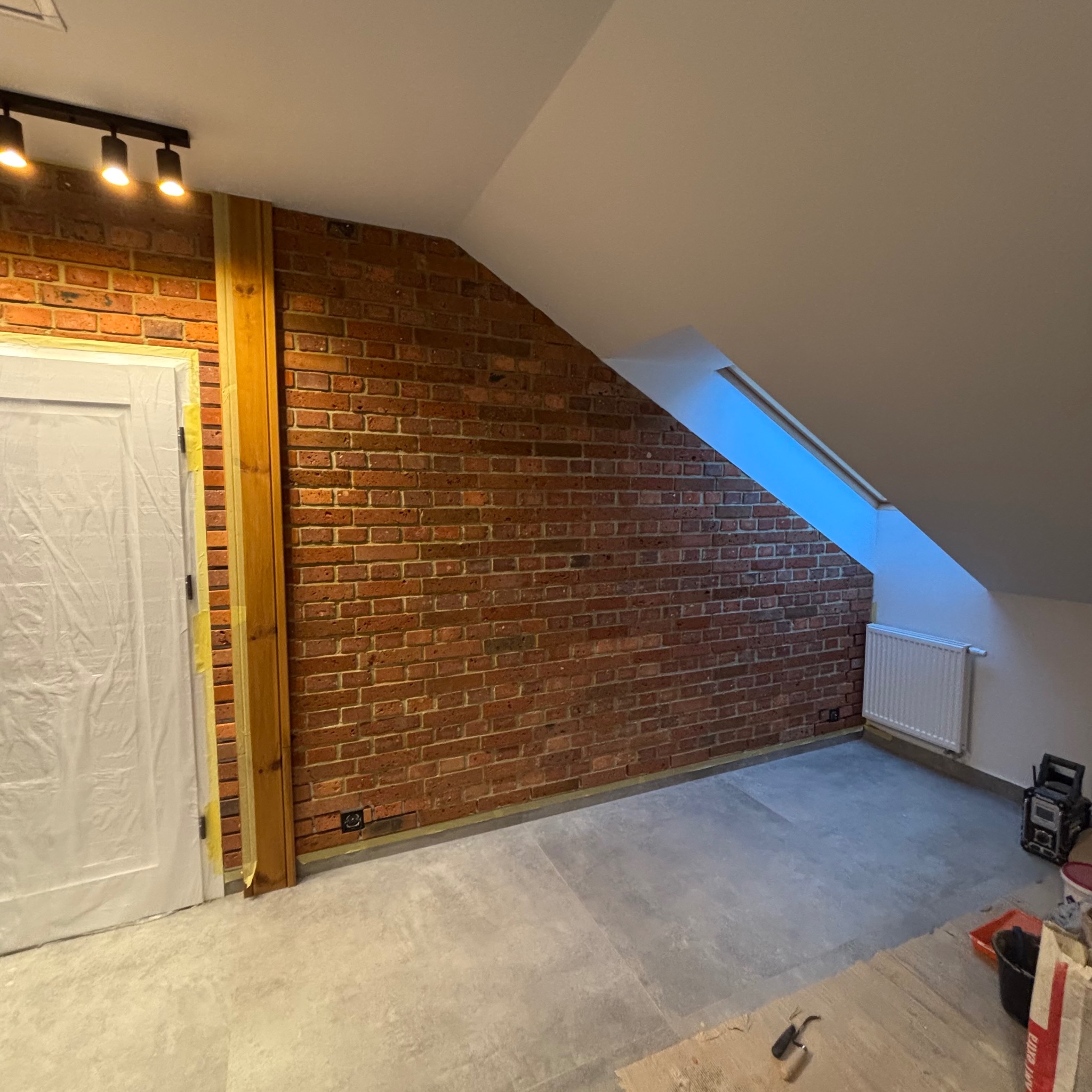 Pokój na poddaszu z ceglaną ścianą, zabezpieczone drzwi folią malarską, widoczne narzędzia i materiały budowlane, okno dachowe w skosie.