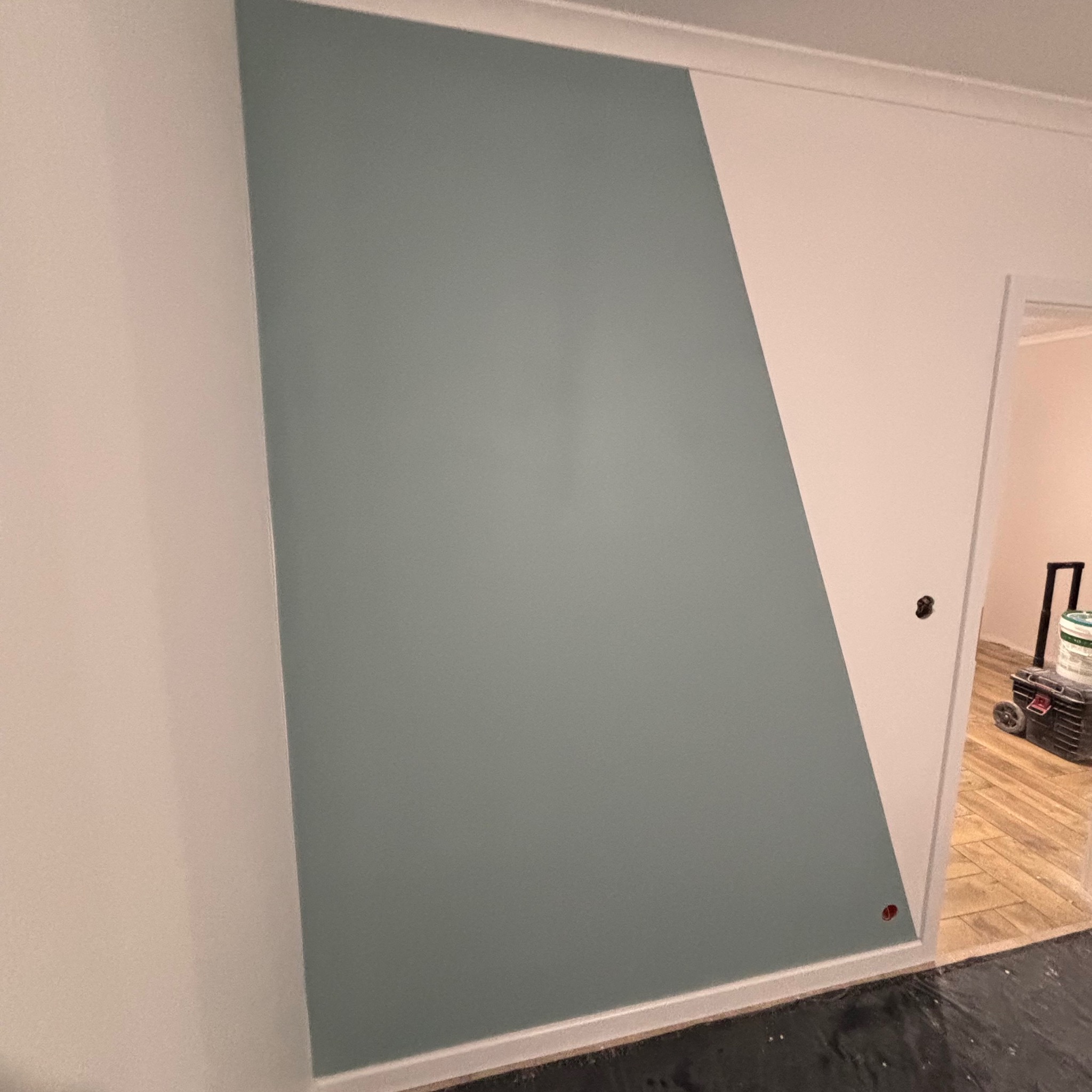 Świeżo pomalowana ściana w Dopiewie, z nietypowym, ukośnym podziałem na kolor szarozielony i biały, widoczny fragment podłogi zabezpieczonej folią malarską oraz skrzynka z narzędziami w tle.