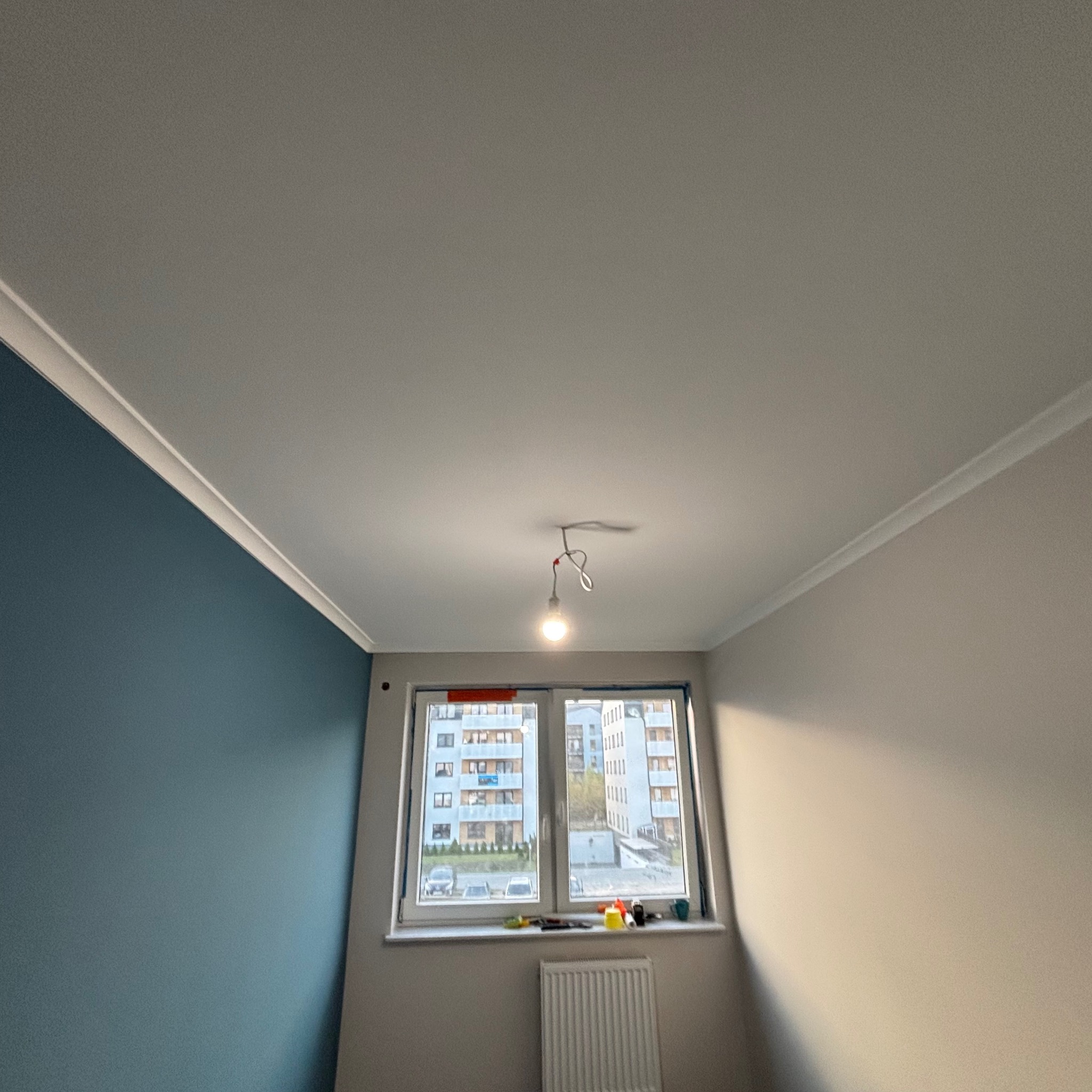 Pokój po remoncie: niebieska ściana kontrastuje z beżową, biały sufit z dekoracyjną listwą, okno z widokiem na budynek mieszkalny, tymczasowa żarówka na kablu.