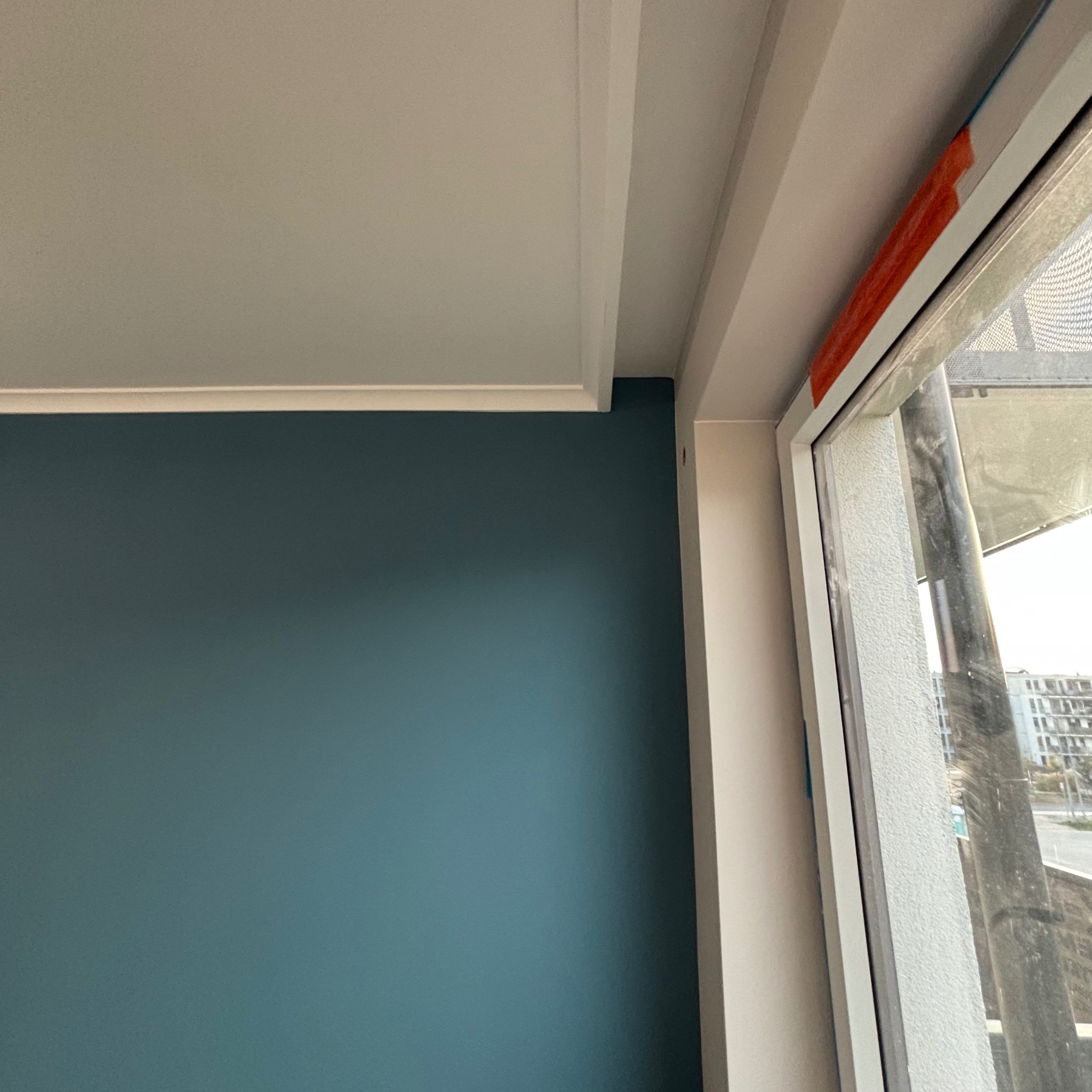 Turkusowa ściana z białym sufitem i listwą przypodłogową, widoczne okno z pomarańczową taśmą zabezpieczającą.