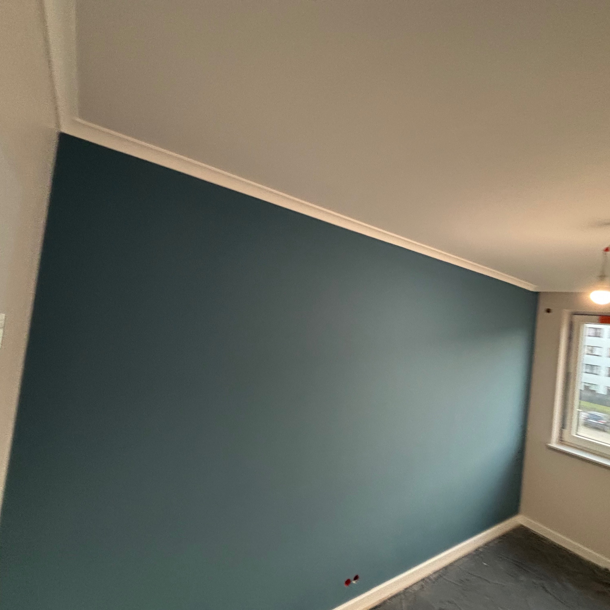Świeżo pomalowana ściana w odcieniu ciemnego turkusu z białym listwą przypodłogową i sufitową, widoczne gniazdka elektryczne bez osłonek, fragment okna z widokiem na zewnątrz.