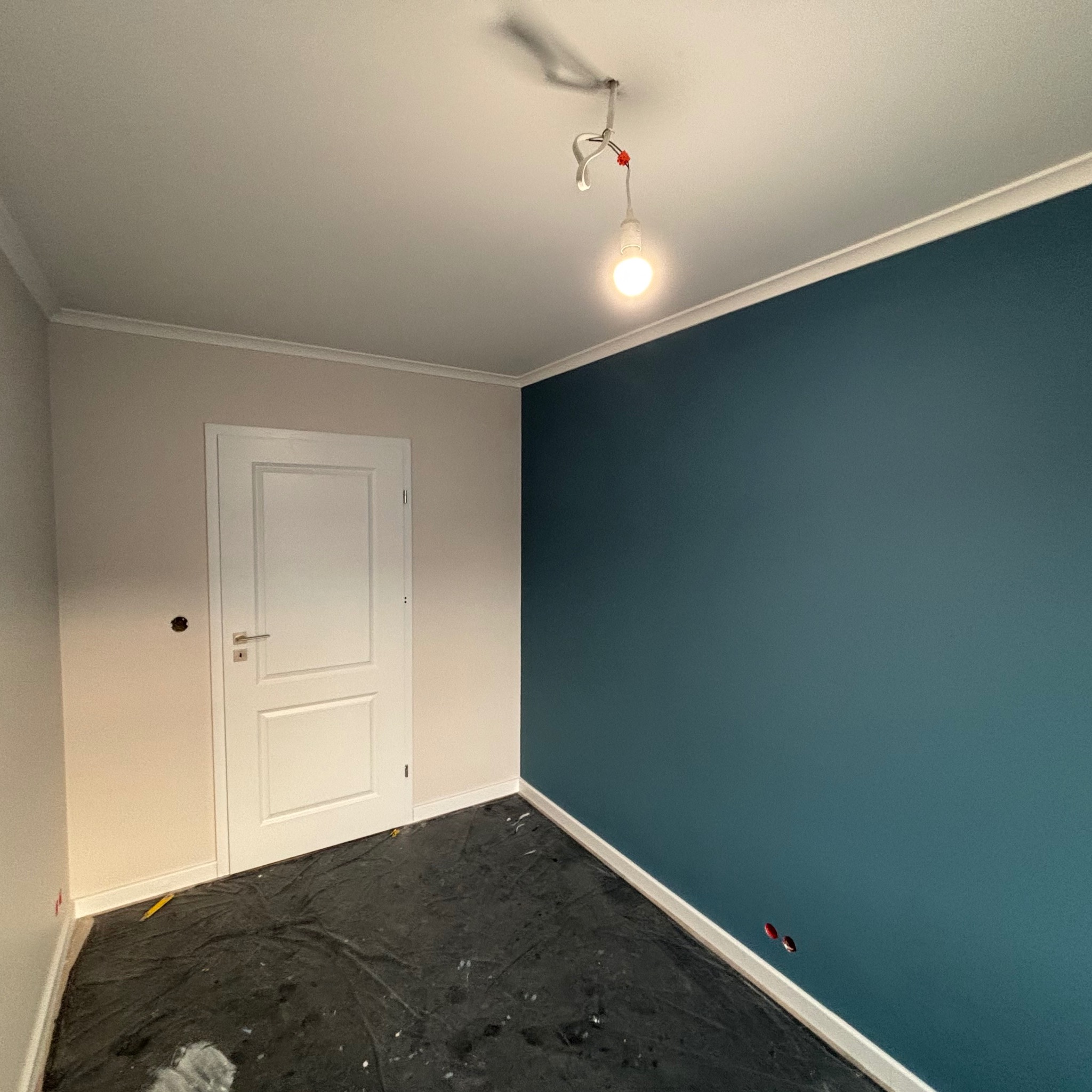 Pokój po malowaniu ścian na dwa kolory: beżowy i ciemny turkus, z białymi listwami przypodłogowymi i osłoniętą folią podłogą, widoczna goła żarówka na suficie.