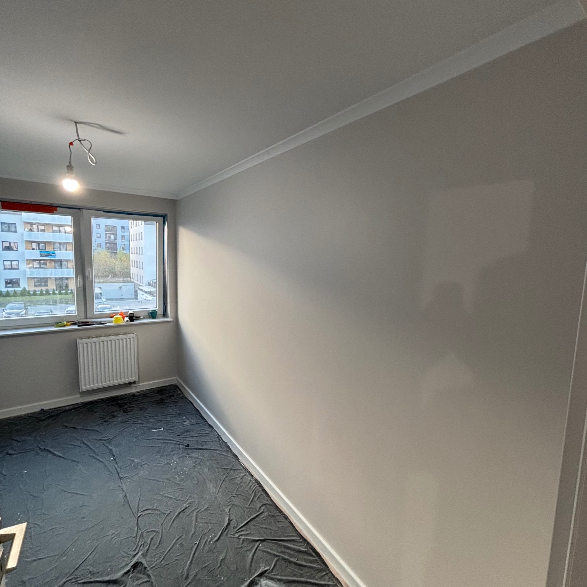 Pokój po malowaniu ścian na szaro, podłoga zabezpieczona folią malarską, widoczne narzędzia malarskie na parapecie, surowa żarówka na kablu.