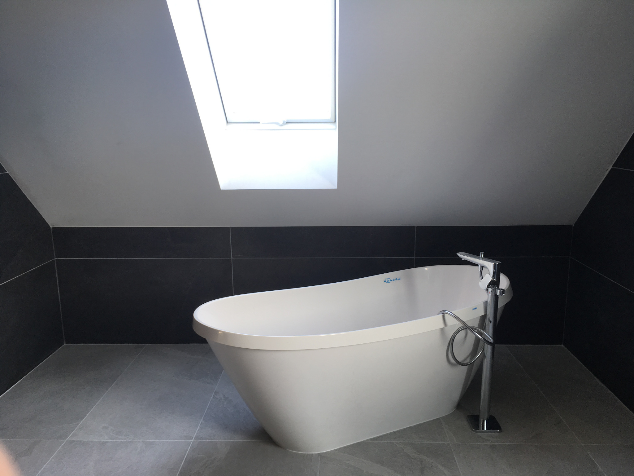 Nowoczesna biała wanna wolnostojąca z chromowaną armaturą na tle szarej płytki podłogowej i ciemnych płytek ściennych w łazience na poddaszu z oknem dachowym.