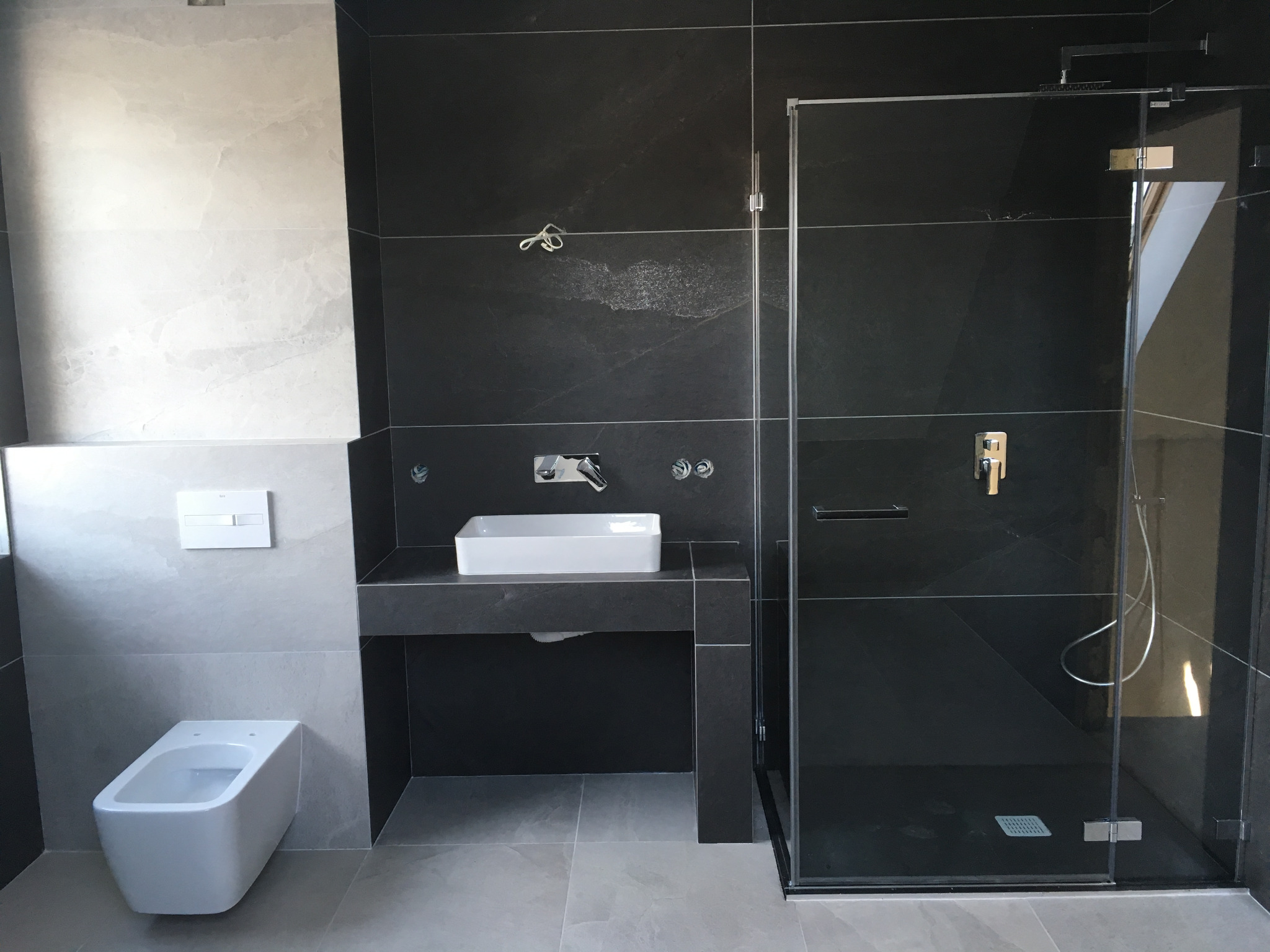 Nowoczesna łazienka z czarnymi płytkami na ścianach, białą ceramiką sanitarną i kabiną prysznicową ze szklanymi drzwiami, minimalistyczna armatura, szare płytki podłogowe.