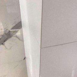 Daniel P - Połączenie białych i marmurowych płytek ściennych w narożniku łazienki, widoczne fugi i fragment drewnopodobnej listwy przypodłogowej.