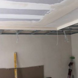 Daniel P - Konstrukcja sufitu podwieszanego z widocznymi profilami metalowymi i przewodami elektrycznymi, częściowo pokryta płytami gipsowo-kartonowymi i warstwą szpachli, w tle pionowa poziomica oparta...