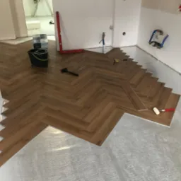 Układanie paneli podłogowych w jodełkę na podkładzie izolacyjnym, widoczne narzędzia: młotek, miara, poziomica, ołówek i wiadro z resztkami.