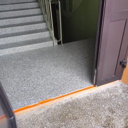 Daniel P - Schody i podłoga w klatce schodowej wykończone szarym lastryko. Widoczne brązowe drzwi i fragment metalowej barierki. Pomarańczowy próg.