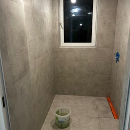 Daniel P - Wykończona łazienka z szarymi płytkami, widoczne okno i wiadro. Minimalistyczny design, surowe oświetlenie. Częściowo widoczna toaleta i pomarańczowa listwa.