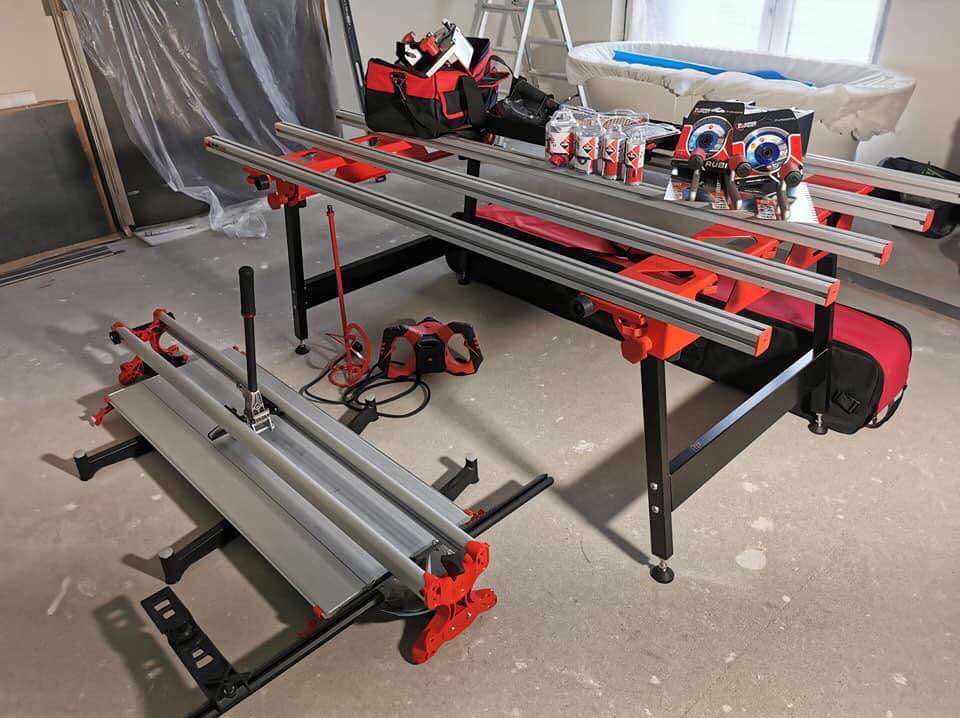 Dwie przecinarki do płytek Rubi, jedna na stole roboczym, druga na podłodze, w tle torba z narzędziami, pojemniki z klejem i drabina.