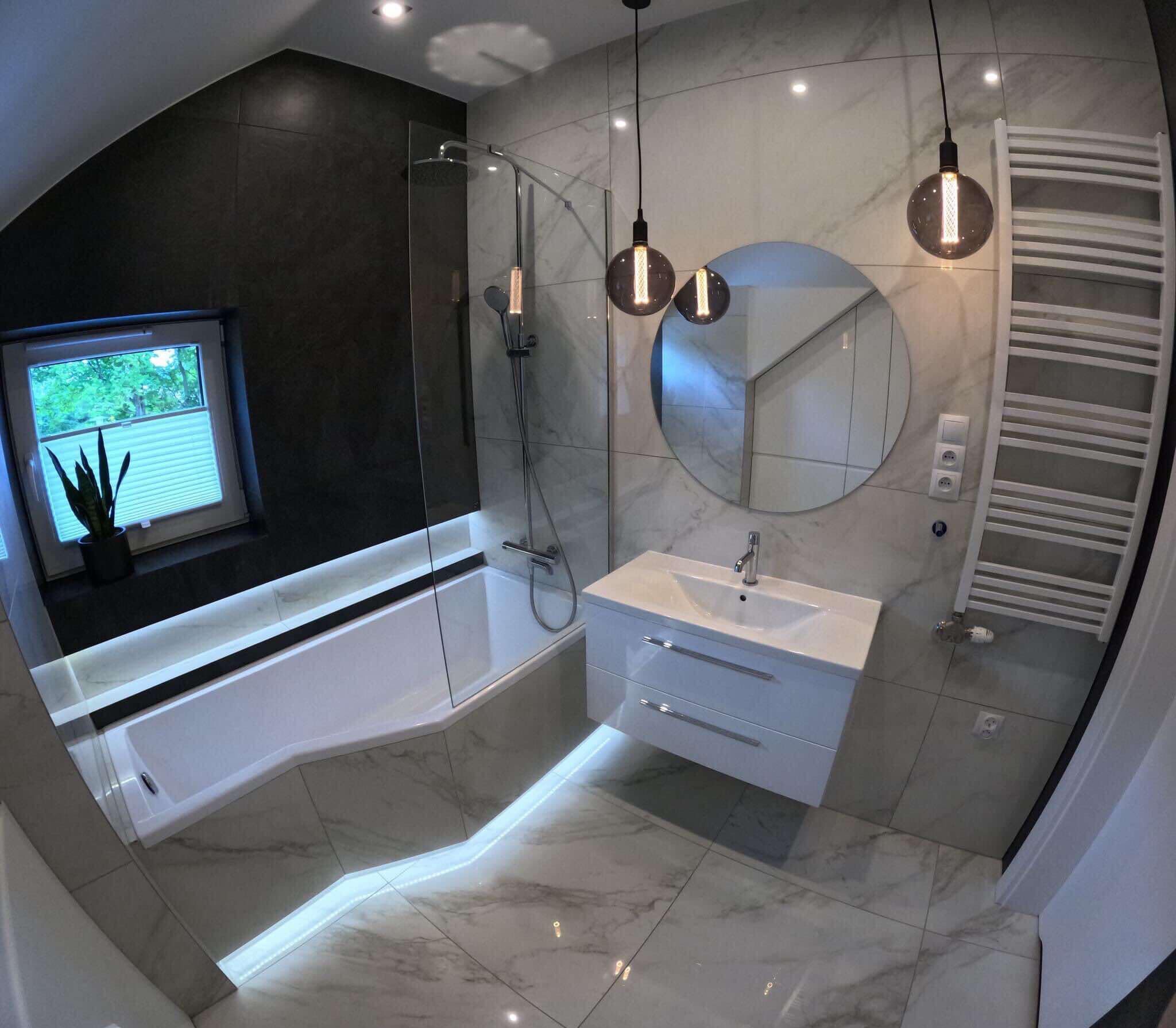 Nowoczesna łazienka z czarno-białymi płytkami marmurowymi, podświetlaną wanną, okrągłym lustrem i wiszącymi lampami z żarówkami Edisona.