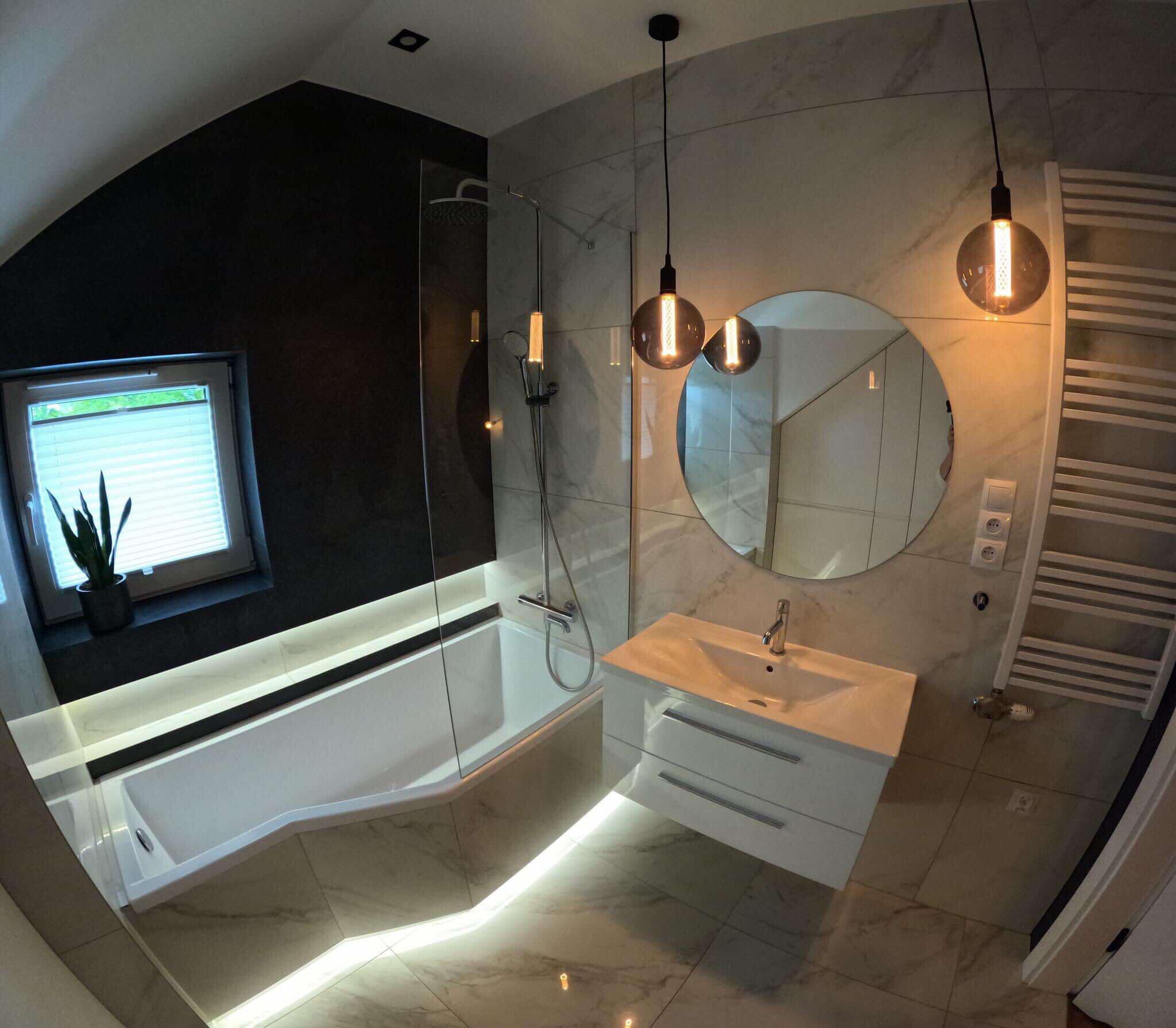 Nowoczesna łazienka z podświetlaną wanną, szklanym parawanem nawannowym, okrągłym lustrem i wiszącymi lampami z żarówkami Edisona, wykończona marmurowymi płytkami.
