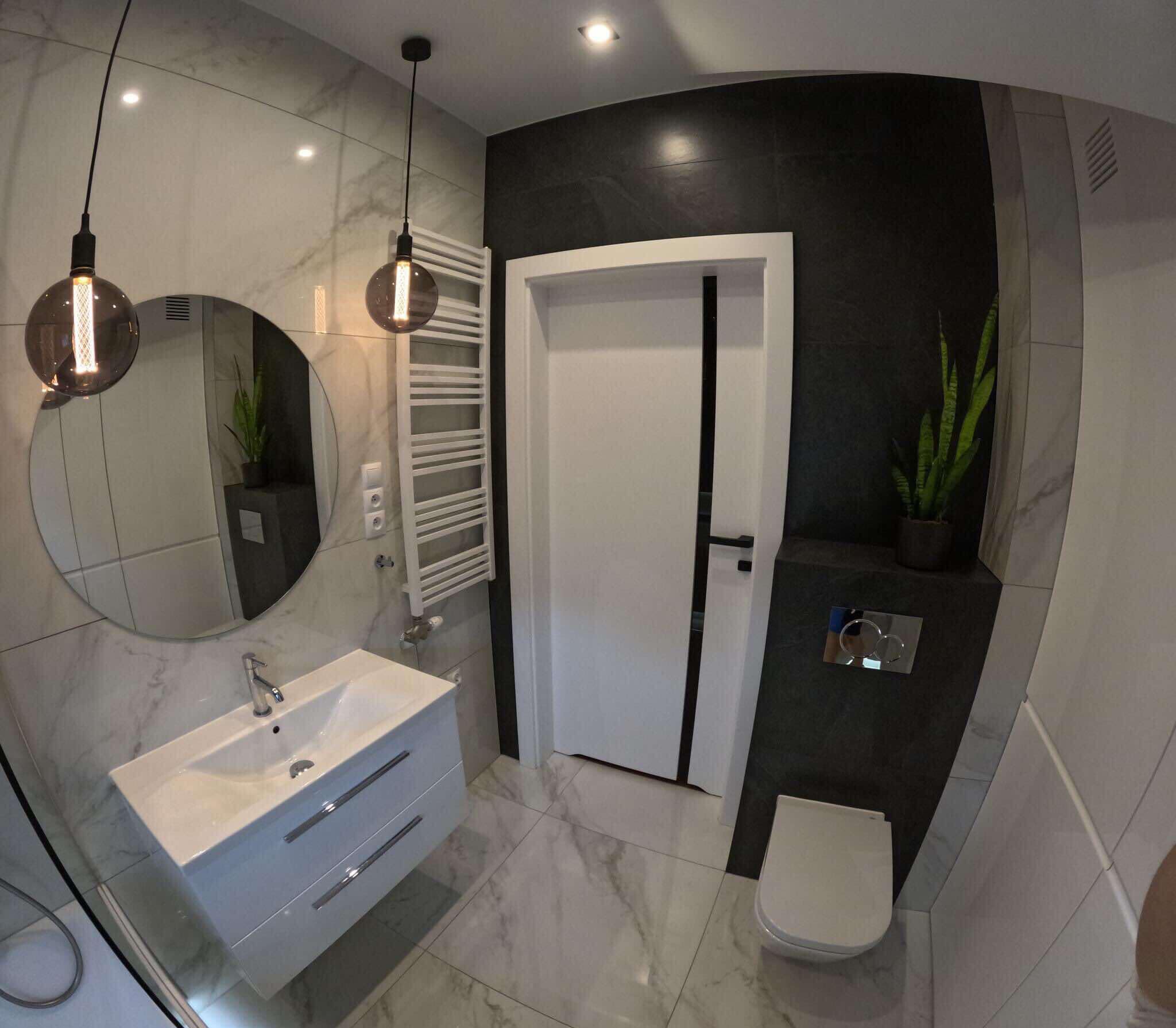 Nowoczesna łazienka z marmurowymi płytkami, okrągłym lustrem podświetlonym dekoracyjną żarówką, wiszącą szafką z umywalką, grzejnikiem drabinkowym i toaletą w zabudowie.