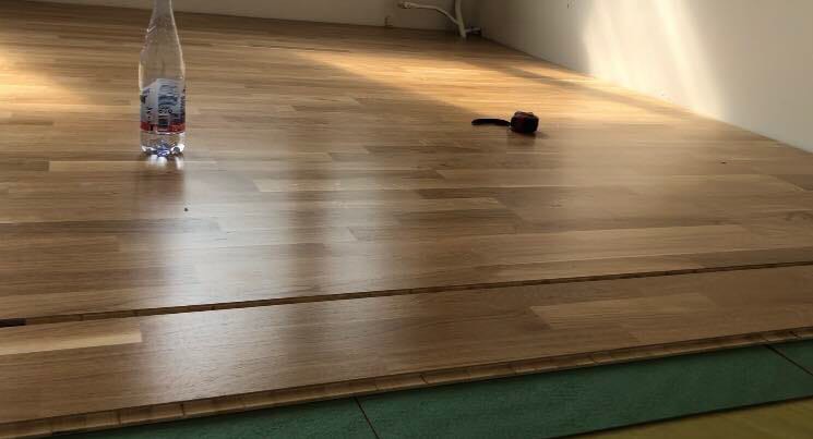 Układanie drewnianych paneli podłogowych, widoczna butelka wody i miarka, podkład w kolorze zielonym.