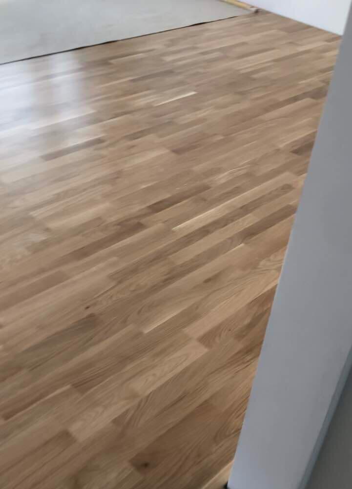 Świeżo ułożona, lakierowana podłoga z paneli drewnianych w odcieniach jasnego brązu, widoczna w perspektywie od rogu pomieszczenia, z fragmentem szarej ściany w prawym dolnym rogu.