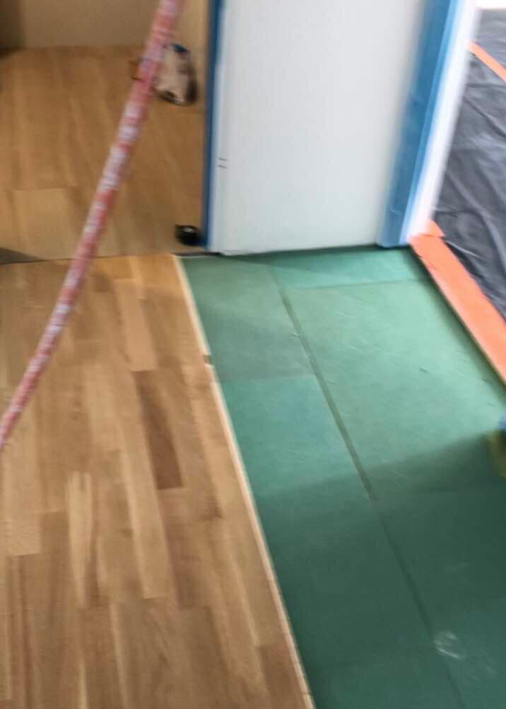 Układanie paneli podłogowych na zielonym podkładzie, widoczny fragment ściany z niebieską listwą przypodłogową.