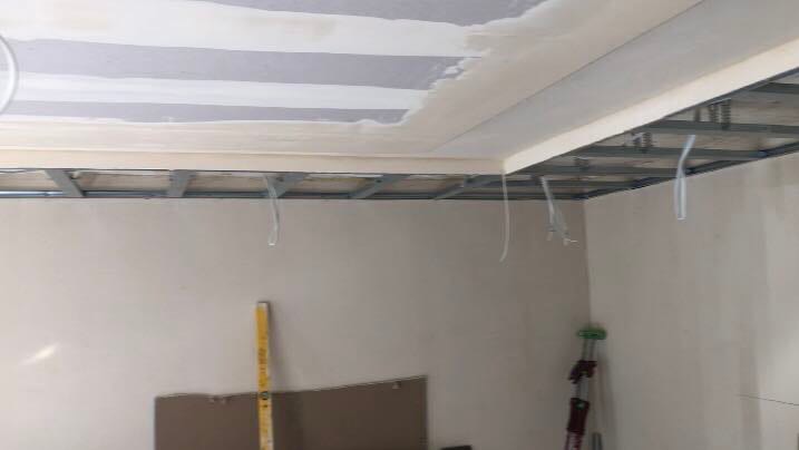 Konstrukcja sufitu podwieszanego z widocznymi profilami metalowymi i przewodami elektrycznymi, częściowo pokryta płytami gipsowo-kartonowymi i warstwą szpachli, w tle pionowa poziomica oparta...