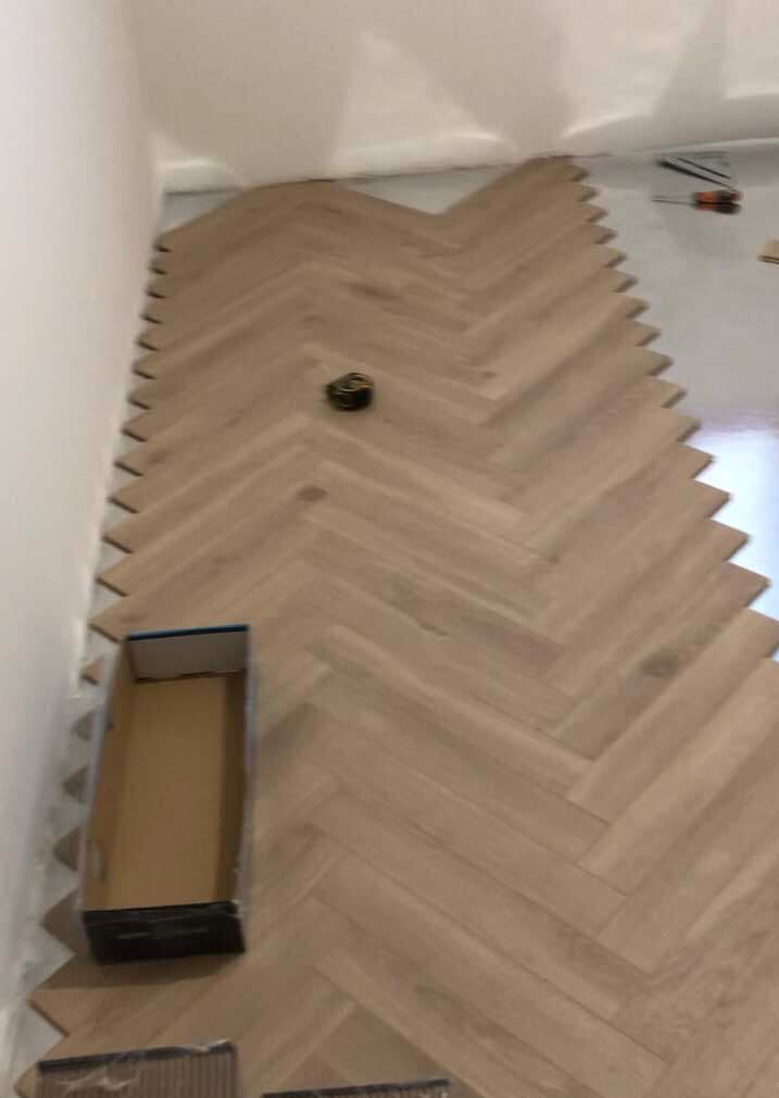 Układanie jasnych paneli podłogowych w jodełkę, widoczna pusta kartonowa skrzynka i narzędzia w tle.