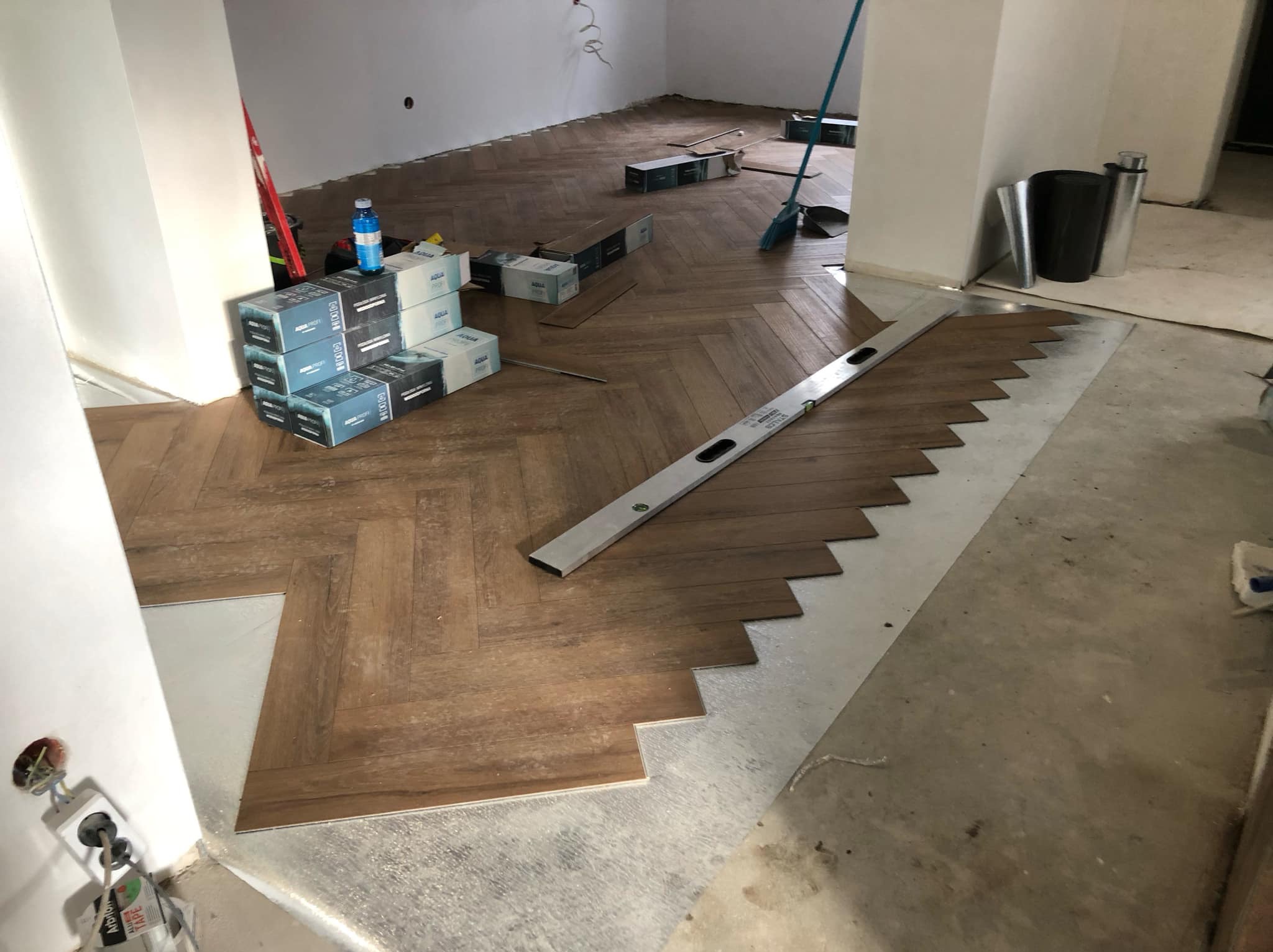 Montaż parkietu w jodełkę: widoczny fragment ułożonej podłogi, poziomica, paczki paneli i przygotowane podłoże z folią izolacyjną.