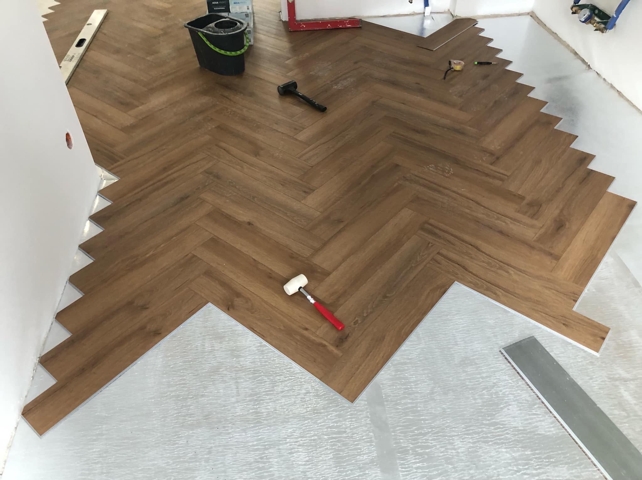 Układanie paneli podłogowych w jodełkę; widoczne narzędzia: gumowy młotek z czerwoną rączką, poziomica, wiadro i podkład izolacyjny.