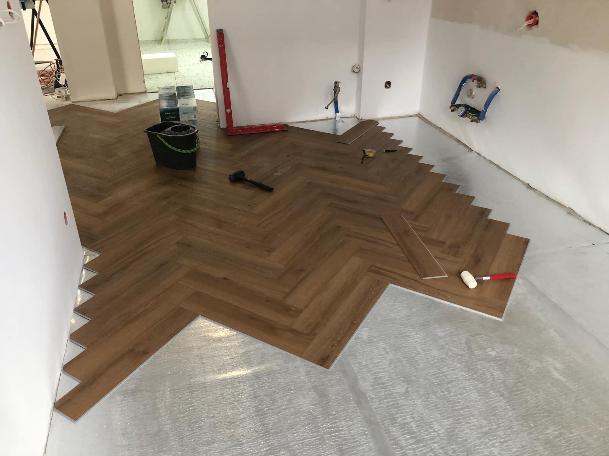 Układanie paneli podłogowych w jodełkę na podkładzie izolacyjnym, widoczne narzędzia: młotek, miara, poziomica, ołówek i wiadro z resztkami.