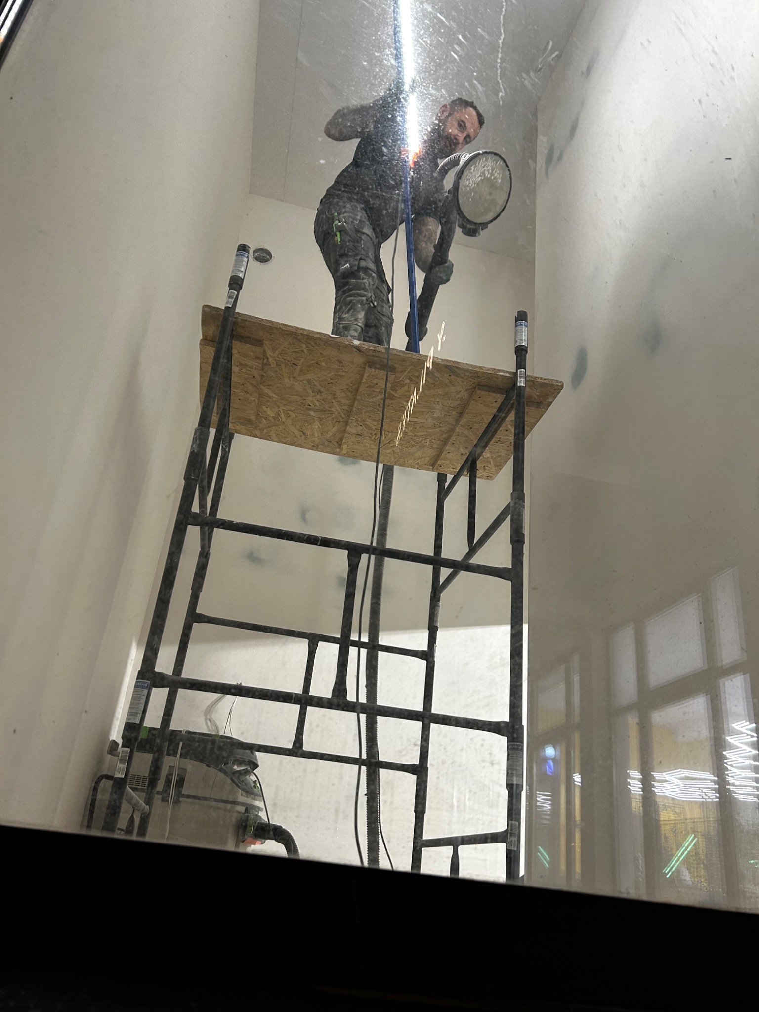 Pracownik na rusztowaniu szlifuje ścianę w pomieszczeniu, oświetlając ją lampą. Widoczny odkurzacz przemysłowy. Przygotowanie do malowania.