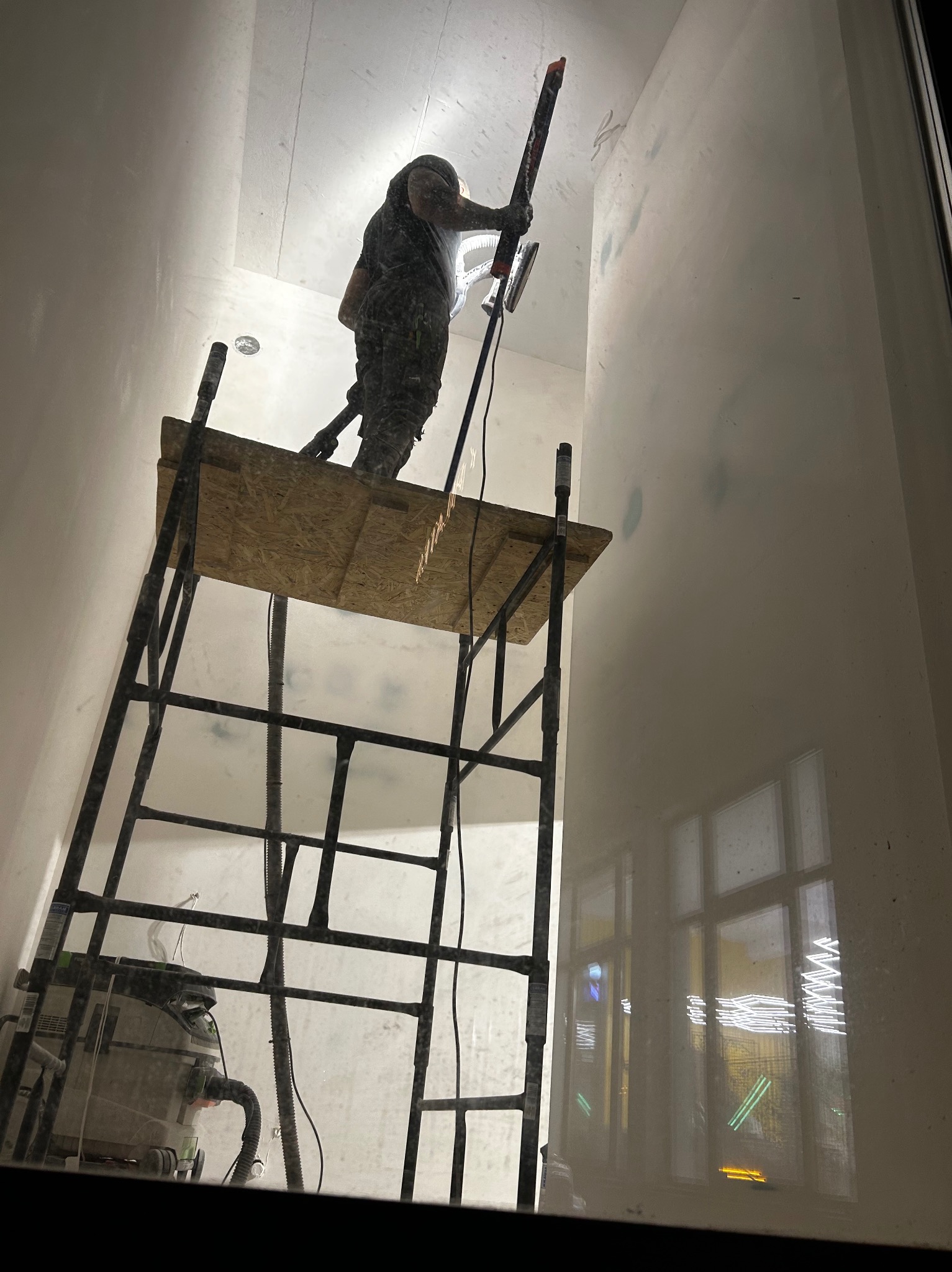 Pracownik na rusztowaniu szlifuje ścianę w pomieszczeniu. Widoczny odkurzacz przemysłowy i okno z odbiciami świateł. Praca w trakcie szpachlowania ścian.