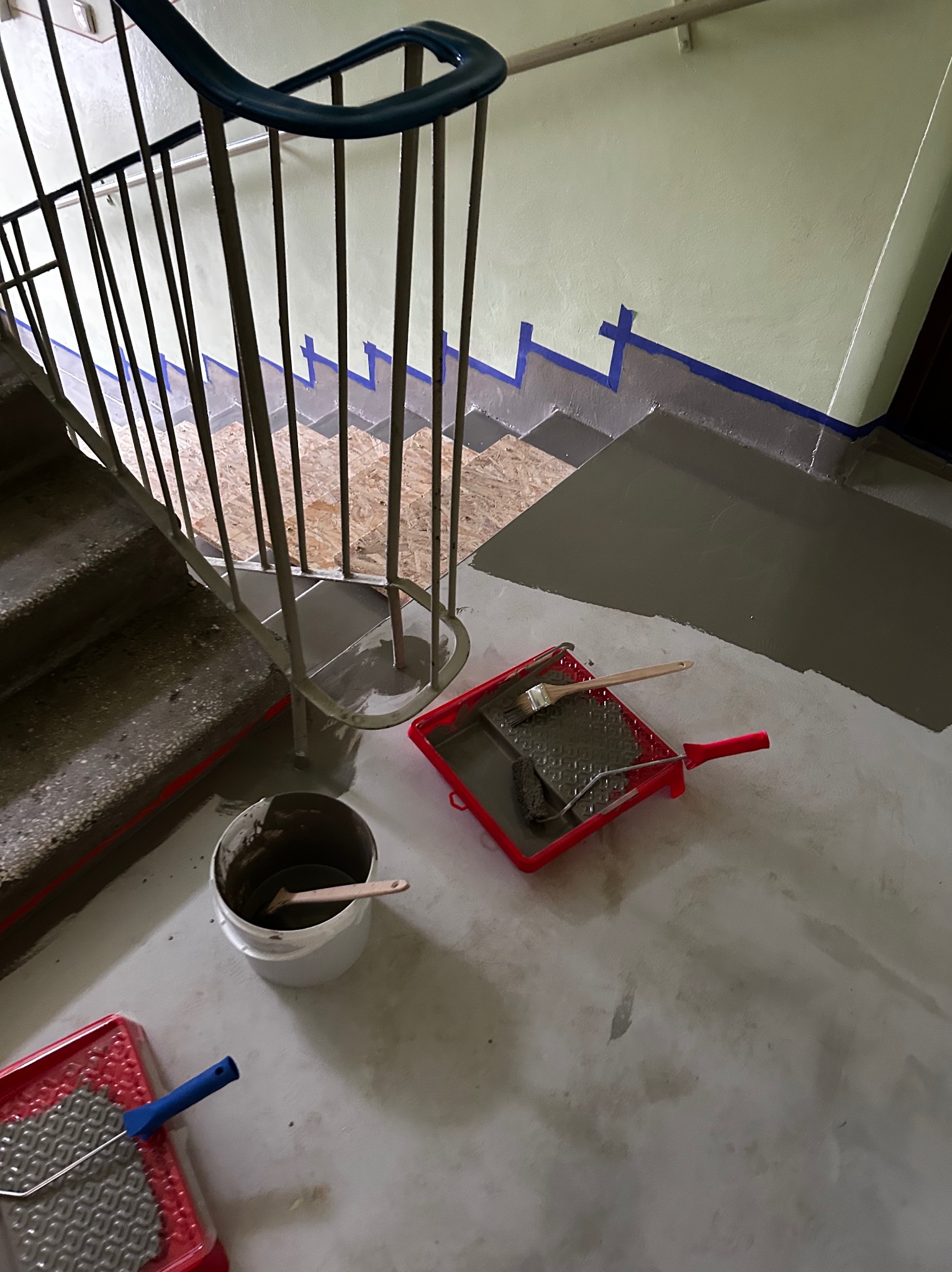 Malowanie klatki schodowej w trakcie prac: pomalowane ściany i podłoga, widoczne narzędzia malarskie, taśma malarska zabezpieczająca krawędzie.