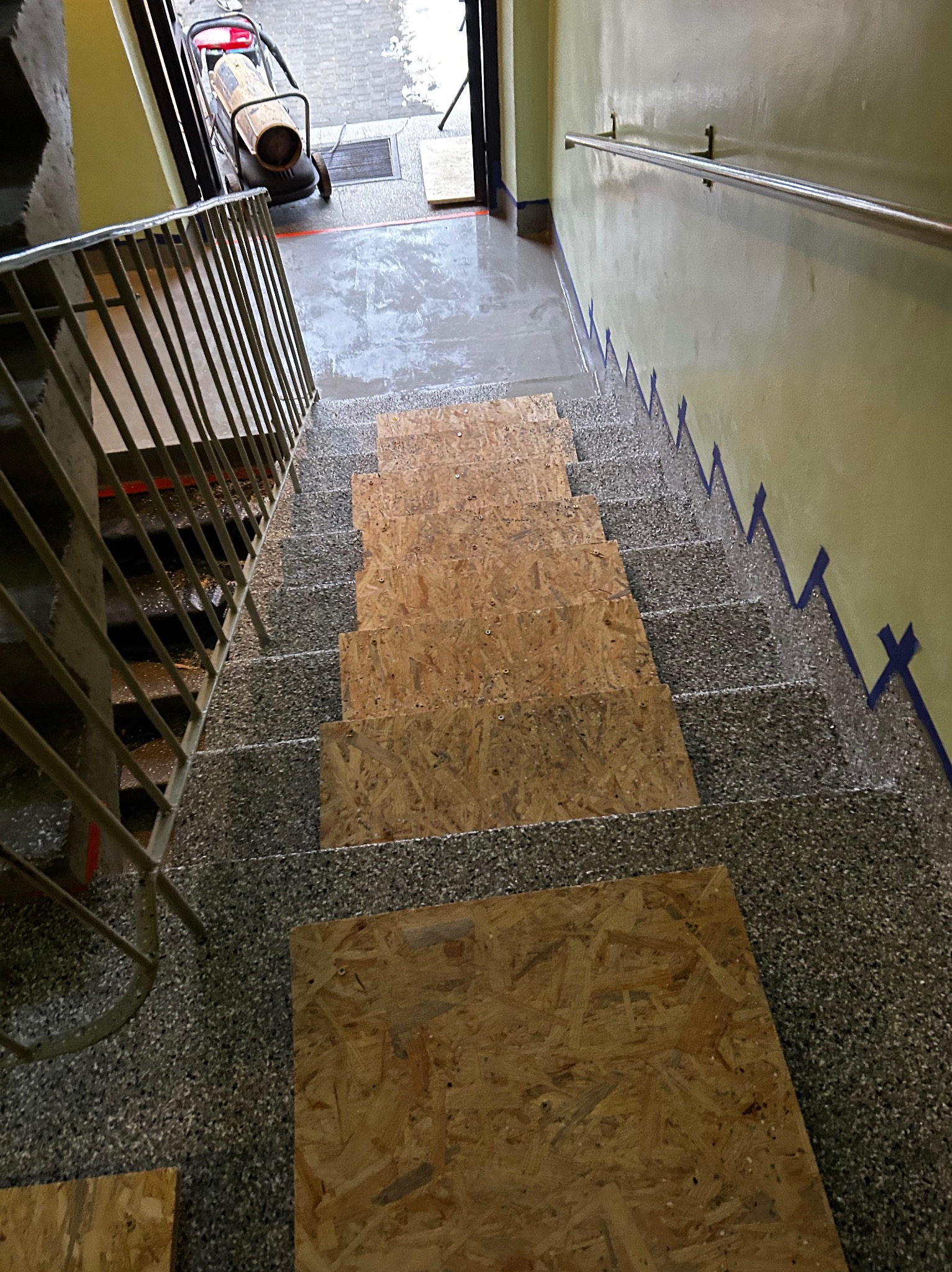 Montaż płyt OSB na schodach w klatce schodowej. Zabezpieczenie schodów na czas remontu. Widoczne taśmy malarskie na ścianach i grzejnik budowlany.